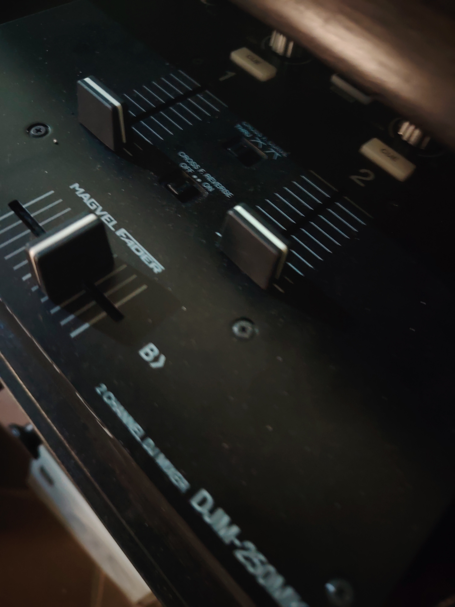 A dark, close-up, slightly angled shot captures the top panel of a black DJ mixer, likely in an indoor, dimly lit environment. The focus is on the central and right sections of the mixer's control surface.
The primary objects visible are several faders and control switches. A prominent black fader with a light grey top is central, labeled "MAGVEL FADER," indicating it's a high-quality crossfader. To its right are two vertical channel faders, each associated with white markings indicating levels and small white "CUE" buttons. The upper parts of these channel faders are partially obscured, but a larger white fader cap is visible on the upper right.
Below the main crossfader, a small switch is labeled "CROSS F. REVERSE" with "OFF" and "ON" positions, currently set to "ON." Other visible text includes "1" and "2" marking the channels, "THRU XA," and "B>" near the crossfader. Along the bottom edge, part of the model name "2 CHANNEL DJ MIXER DJM-250MK" is legible. The surface of the mixer shows some faint dust particles and minor wear, consistent with use. The lighting casts subtle reflections on the faders and highlights the embossed text. No people are present, and no specific activity is taking place, only the equipment is showcased.