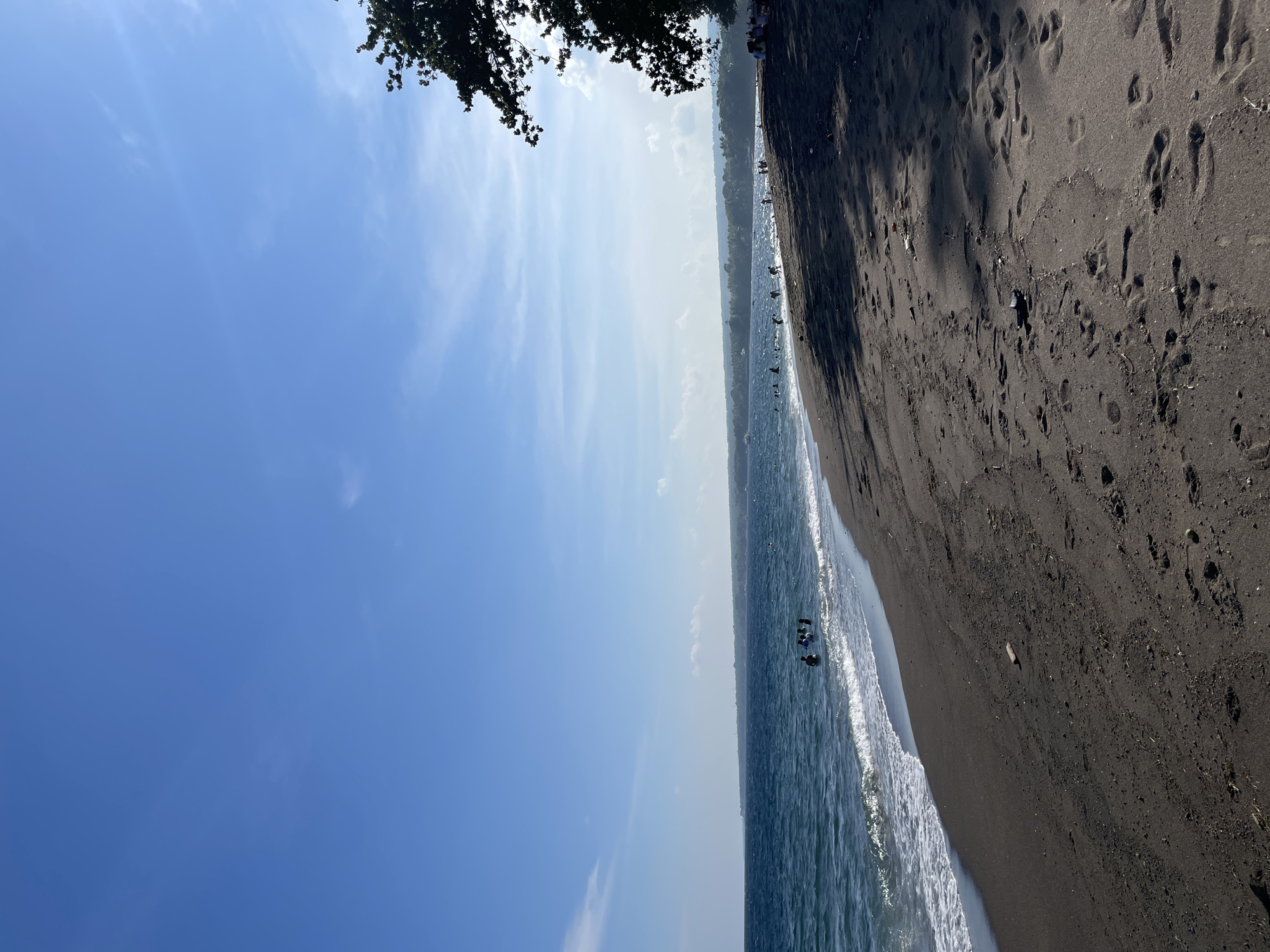 This image captures a serene beach scene on a clear, sunny day in Dauin, Philippines. The foreground is dominated by dark, volcanic sand, showing numerous footprints and a few scattered pieces of driftwood and debris. Gentle waves roll onto the shore, creating a white foamy line along the water's edge.
In the middle ground, several people are visible in the ocean, some appearing to be swimming or wading. The water is a deep blue, reflecting the clear sky above. A distant coastline stretches across the horizon, lined with lush green vegetation and hinting at more populated areas further inland.
To the right, the silhouette of a large tree branches out, casting shadows across the sand, suggesting the sun is positioned in the upper left of the frame. The sky is a vibrant blue, with a few wispy clouds adding texture. The overall atmosphere is peaceful and inviting, typical of a tropical beach destination.
