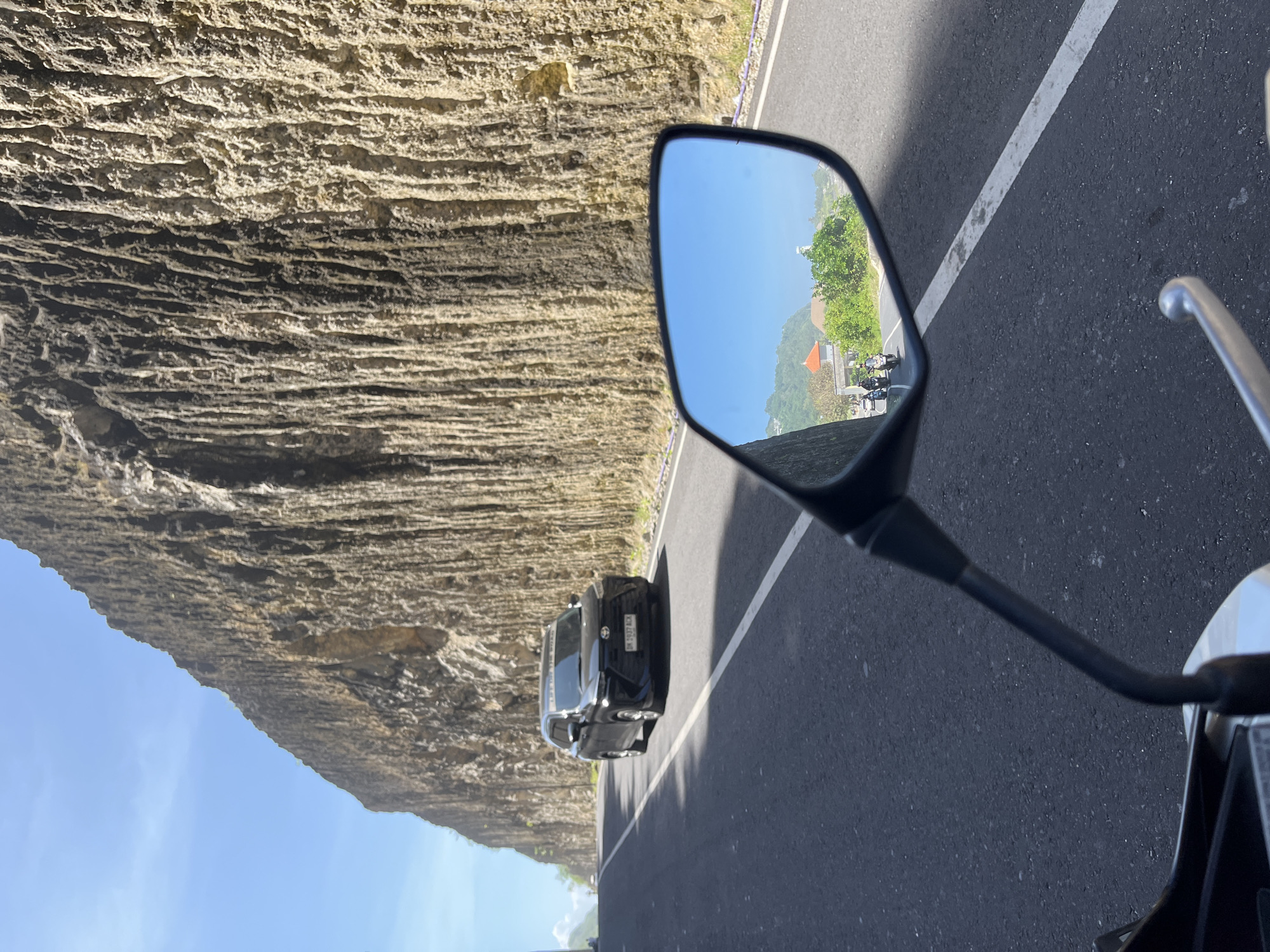 The image shows a view from a motorcycle on a road in Kuta Selatan, Indonesia. A high rock wall runs along the left side of the road. A black car is behind the motorcycle. The motorcycle's side mirror reflects a scene of a road with other motorcycles and a distant building. The sky is blue.