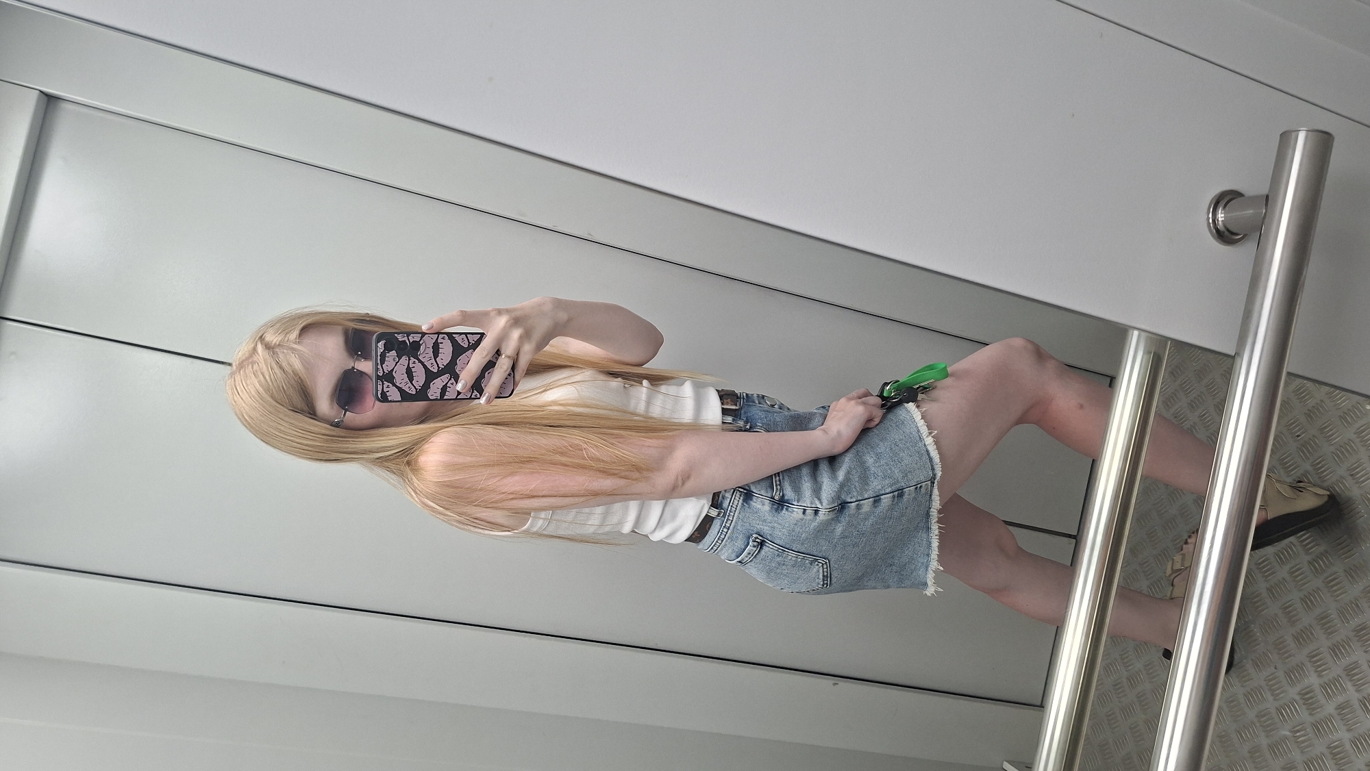 A pale-skinned person with long blonde hair, wearing sunglasses, a white tank top, and a denim skirt, stands in what appears to be an elevator. The person is holding a smartphone with a pink lip-patterned case, taking a selfie. The elevator has a metallic floor with a raised diamond pattern, a silver handrail, and white walls. The location in the city is Unknown, Unknown.