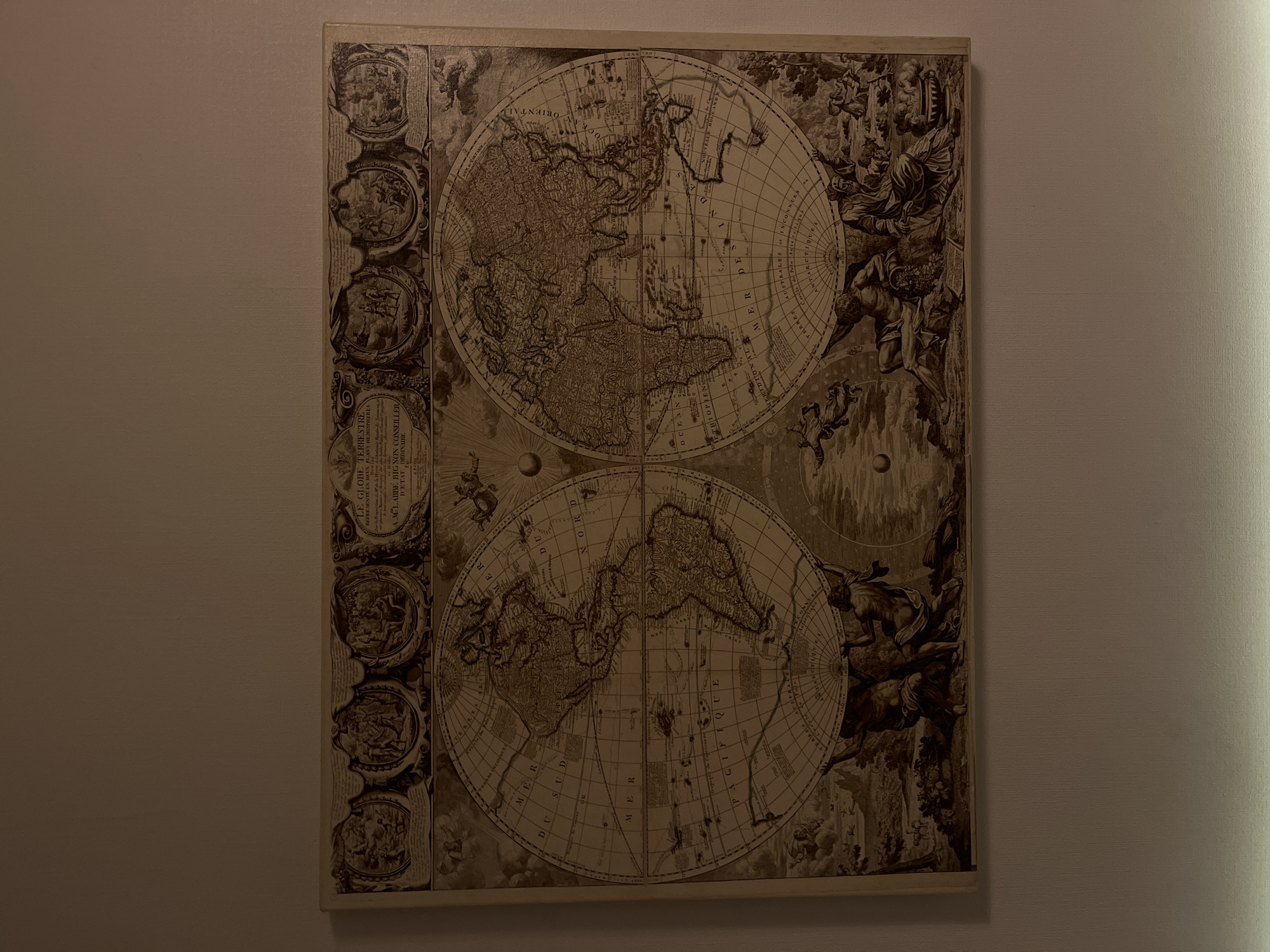 An antique-style map of the world, depicting two hemispheres, is displayed on a light-colored wall. The map is a sepia-toned etching, featuring detailed coastlines, landmasses, and bodies of water. It is framed by decorative borders at the top and bottom. The top border contains several circular vignettes with figures, possibly allegorical or historical. The bottom border depicts various figures, including two powerful male figures holding up the map, surrounded by mythical or classical scenes. The map itself is divided by lines of latitude and longitude and includes labels in what appears to be French. The overall impression is one of historical significance and artistic craftsmanship. The scene is indoors, with the map mounted on a wall in what appears to be a domestic or exhibition setting.