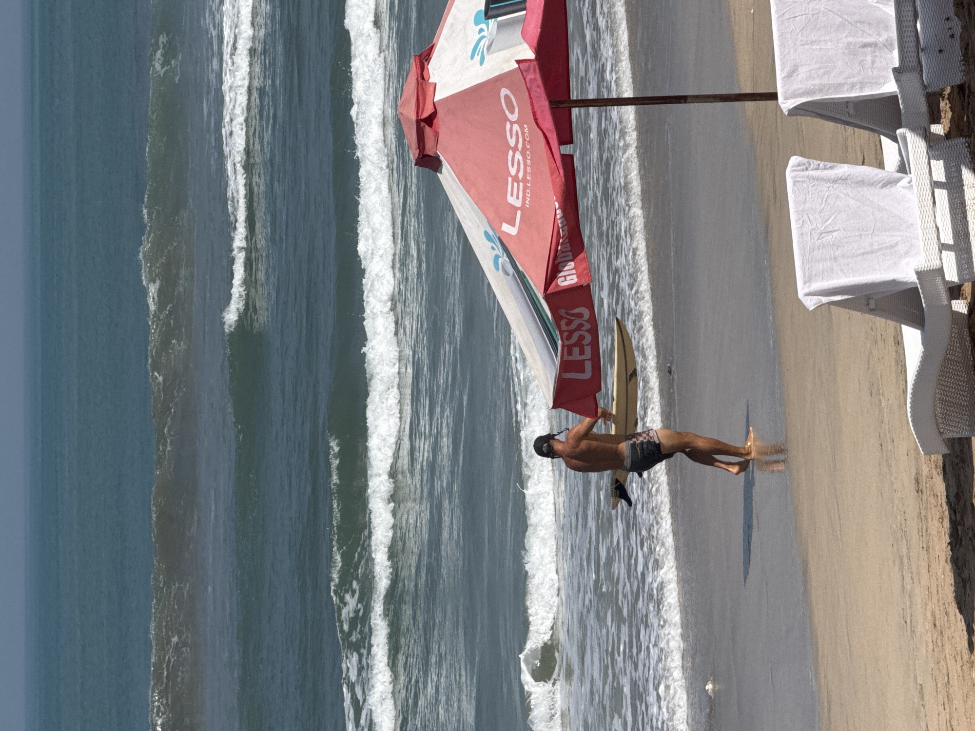 In Kuta, Indonesia, a lone male surfer, wearing a black cap and checkered swim trunks, walks from the ocean towards the shore, carrying a surfboard. He is heading towards a red and white beach umbrella with the word "LESSO" prominently displayed. The umbrella is set up on the sandy beach, and two white lounge chairs are visible in the foreground, draped with white towels. The ocean in the background features moderate waves with white foam crests, indicating typical surfing conditions. The sky is a hazy blue, and the overall atmosphere suggests a sunny day at the beach.