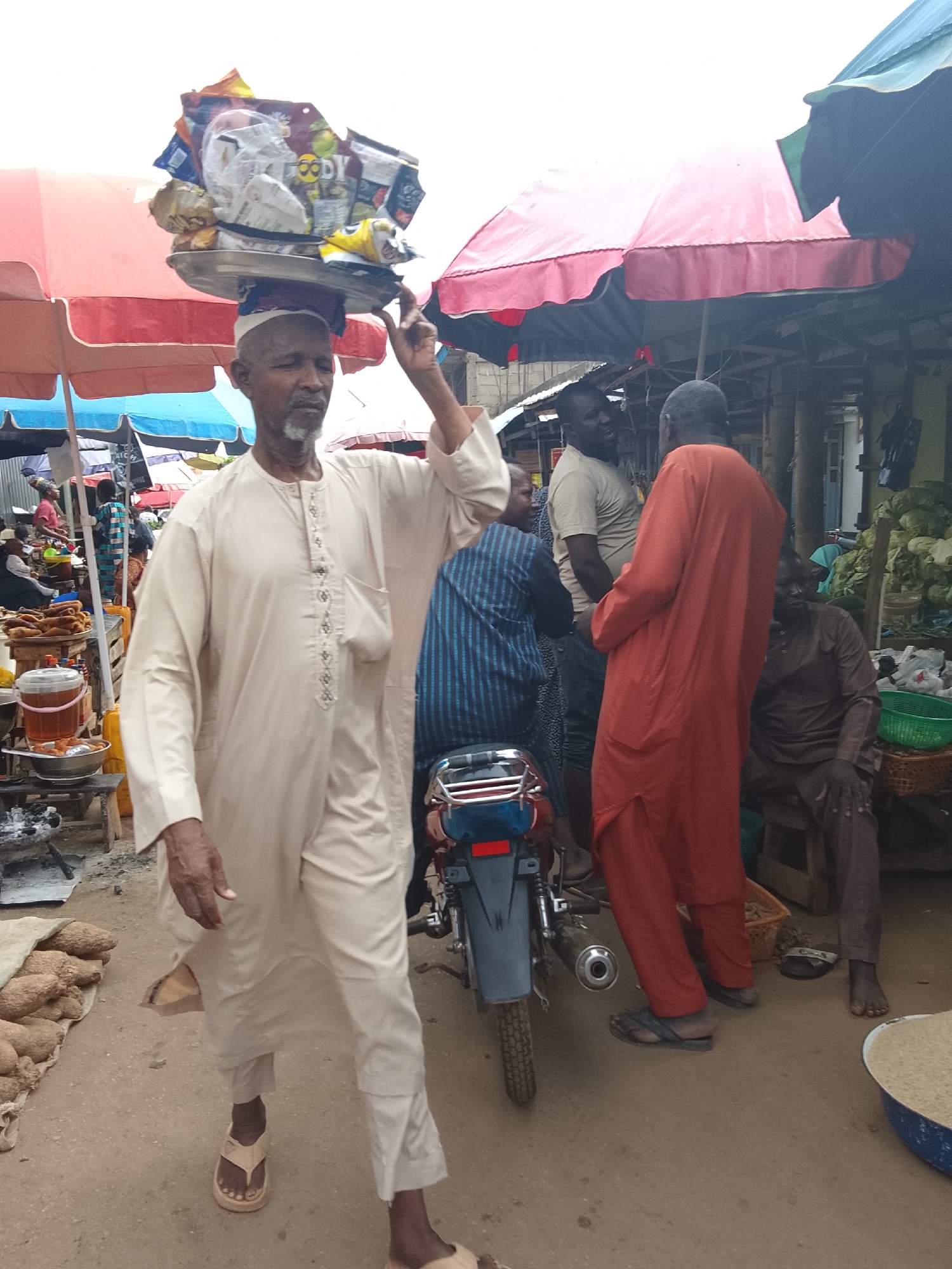 In Akwanga, Nigeria, a busy marketplace scene unfolds. A man in a light-colored kaftan walks forward, carrying a tray of snacks balanced on his head. He wears sandals. Behind him, other individuals are present, including a person in an orange robe and a man in a brown tunic. A motorcycle is parked nearby. Stalls with umbrellas are visible, suggesting a lively trading environment. Several people are engaged in conversation or are simply present within the market. The ground is dirt, and there are sacks of produce and baskets of food items visible, indicating a typical market setting where goods are bought and sold.