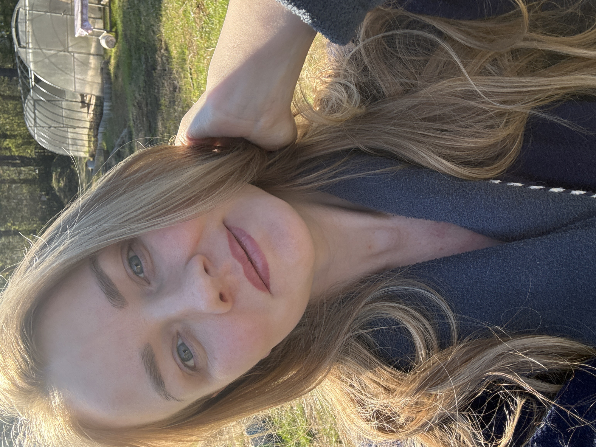 A blonde woman is positioned in the foreground, her head tilted. Her hand is raised and placed behind her head. In the background, to the left, is a greenhouse. The background is grassy. The location appears to be rural and could be consistent with an area outside of Chaikovsky, Russia.