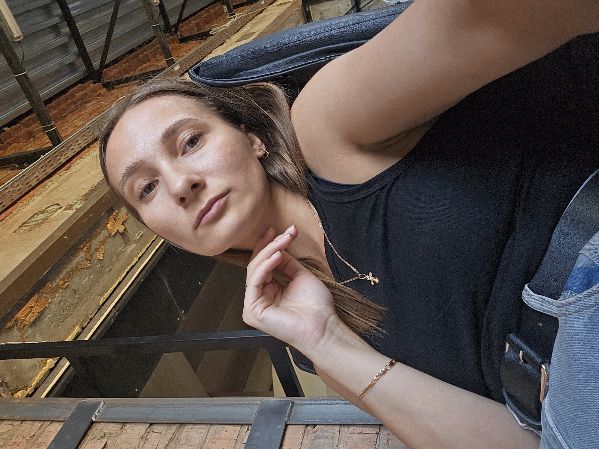 A light-skinned woman with brown hair is captured in a selfie-style photograph, seemingly indoors in a building in Chaikovsky, Russia. She is dressed in a black tank top and denim jacket. A black purse sits on her shoulder. She is wearing a gold chain necklace and a gold bracelet on her left wrist. Her left hand is raised, holding a piece of her hair. The setting appears to be a building under construction or renovation, evidenced by the exposed brick and metal supports overhead. The photo is taken at an unusual angle, looking upwards at the subject.
