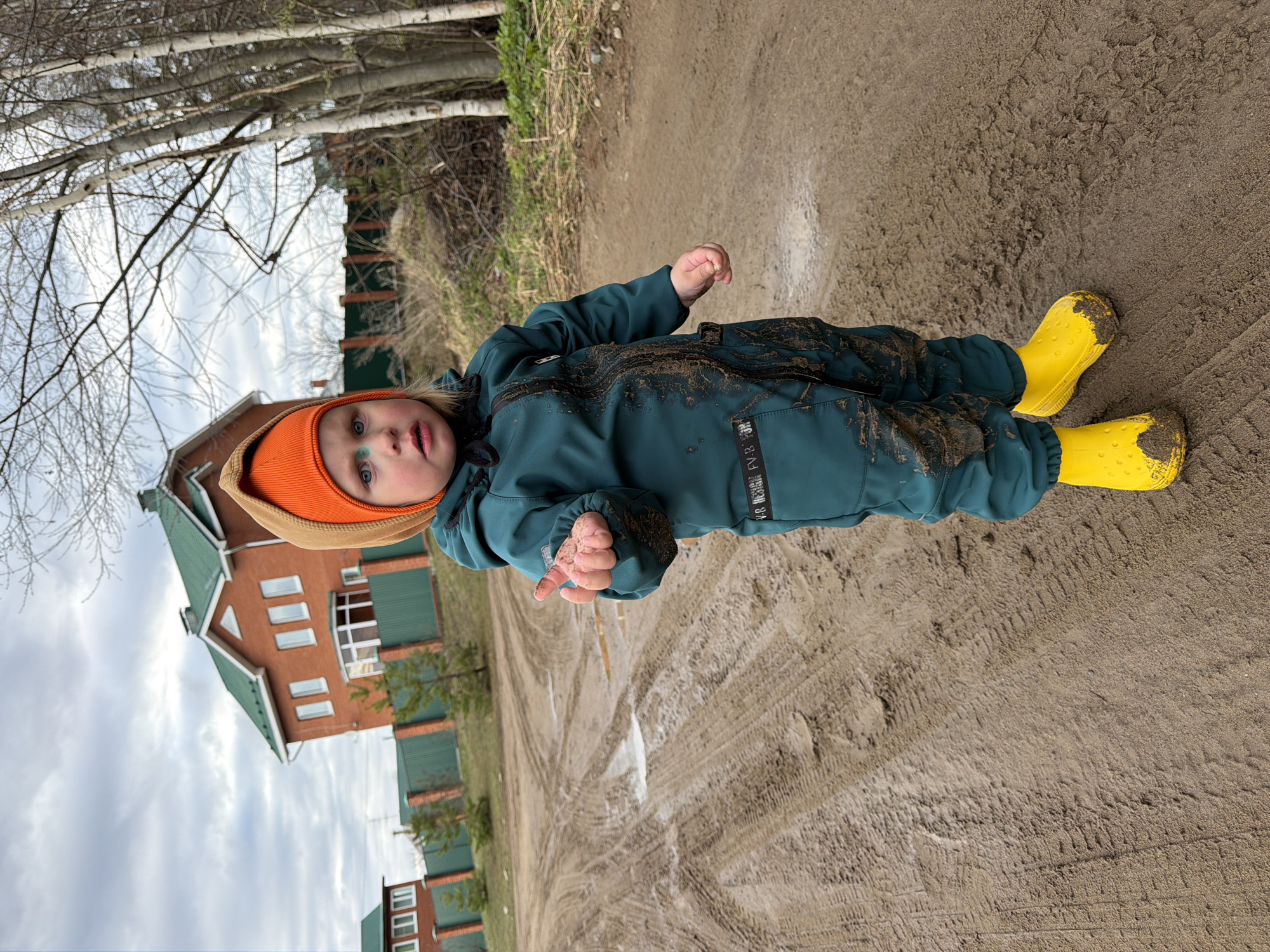 An outdoor, slightly off-angle portrait shows a young child standing on a muddy, unpaved path under an overcast sky. The child, likely a toddler, wears a dark teal waterproof jumpsuit heavily splattered with mud, bright yellow rubber boots also covered in mud, and an orange knit hat with a beige brim. A small blue mark is visible under their left eye. They look directly at the camera with a neutral expression, holding a small amount of dirt or mud in their right hand.
The ground is very wet and muddy, displaying distinct tire tracks. In the background to the left, several tall, bare deciduous trees, possibly birch, stand against the cloudy sky. To the right, a multi-story brick house with a green roof and white windows is partially visible behind a dark green fence. Another building can be seen further in the distance.
The scene suggests a cool, damp day, likely after rain, consistent with a rural or suburban setting in Chaikovsky, Russia. The child appears to be engaging in muddy play or exploration. A white label with "FV-8." and other small text is visible on the left leg of the jumpsuit.
