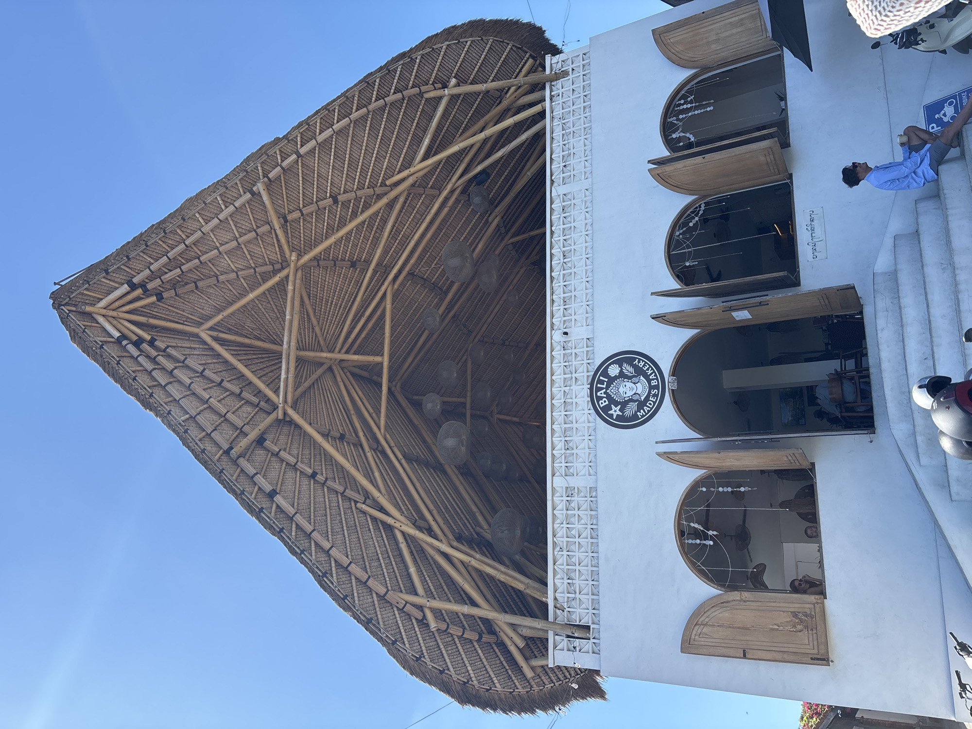 A low-angle, full exterior shot depicts a two-story commercial building identified as "BALI MADE'S BAKERY" in Kuta Selatan, Indonesia, under a clear, bright blue sky. The most prominent feature is the large, conical, thatched roof, constructed from bamboo poles and natural straw/thatch material, forming an open, high-ceilinged upper level. Below the roof, the upper story features a white concrete balustrade with a geometric openwork pattern. Spherical, woven light fixtures hang from the exposed bamboo rafters within the upper roof structure.
The lower story is a white-rendered facade with three arched openings. These openings are fitted with wooden bi-fold shutters, two of which are open, revealing the interior of the bakery. Through the left open window, two individuals are visible, one looking outwards wearing sunglasses. Decorative string lights or ornaments hang within the window arches. Wide, light-colored concrete steps lead up to these entrances.
On the lower right steps, a male individual is seated, wearing a light blue long-sleeved shirt, dark shorts, and sunglasses, holding a cup. A blue sign indicating "P BIKE" (likely parking for motorbikes) is visible near his foot. Partial views of parked motorbikes or scooters and helmets are visible at the base of the steps on the left and right sides. A black umbrella is partially visible on the far right. The scene is brightly lit by natural sunlight.