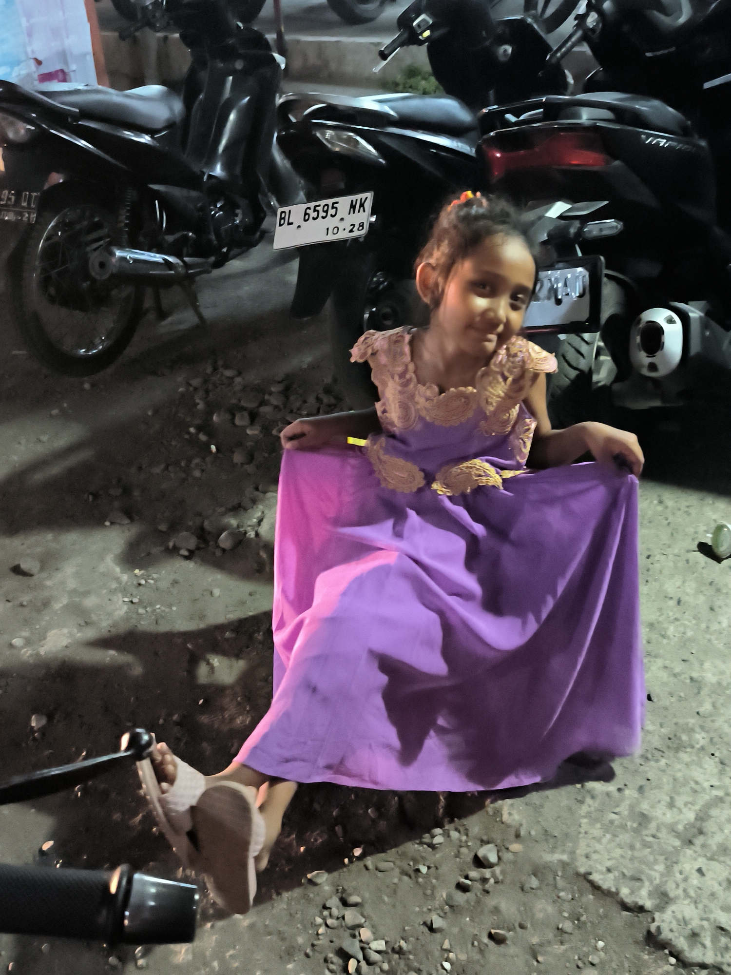 A young girl sits on the ground in Lhokseumawe, Indonesia. She is wearing a floor-length purple dress with gold lace detailing. Behind her are parked motorbikes, one with a license plate reading "BL 6595 NK 10-28". The ground is a mix of pavement and dirt with small rocks. Part of a motorbike handlebar is visible in the foreground. The lighting suggests it is nighttime.