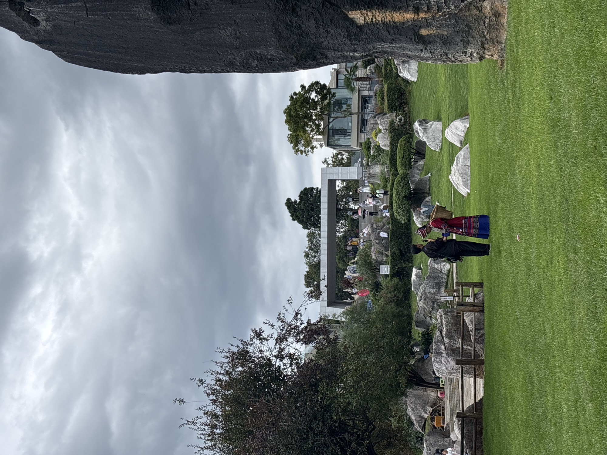 The image captures a scene in the Stone Forest National Park, a geological wonderland located approximately 90 kilometers southeast of Kunming, China. In the foreground, a vast expanse of meticulously manicured green grass stretches across the frame. To the right, a colossal, dark gray rock formation dominates the edge of the image, its textured surface suggesting ancient weathering.
Two individuals stand on the grass in the mid-ground. One, dressed in a black garment, appears to be speaking into a microphone or recording device, facing a second individual. This second person is adorned in a vibrant, traditional red and blue costume, featuring intricate embroidery and patterns. They are carrying a woven basket over their shoulder.
Behind them, a wooden fence delineates an area with scattered, large gray boulders. Further back, a landscape unfolds with more rocky outcrops, manicured bushes, and trees. A modern, multi-story building with large glass windows is visible amongst the greenery, and a wide, elevated walkway with people on it is set against a backdrop of a cloudy, overcast sky. The overall impression is that of a cultural performance or demonstration taking place within the park's unique natural setting.