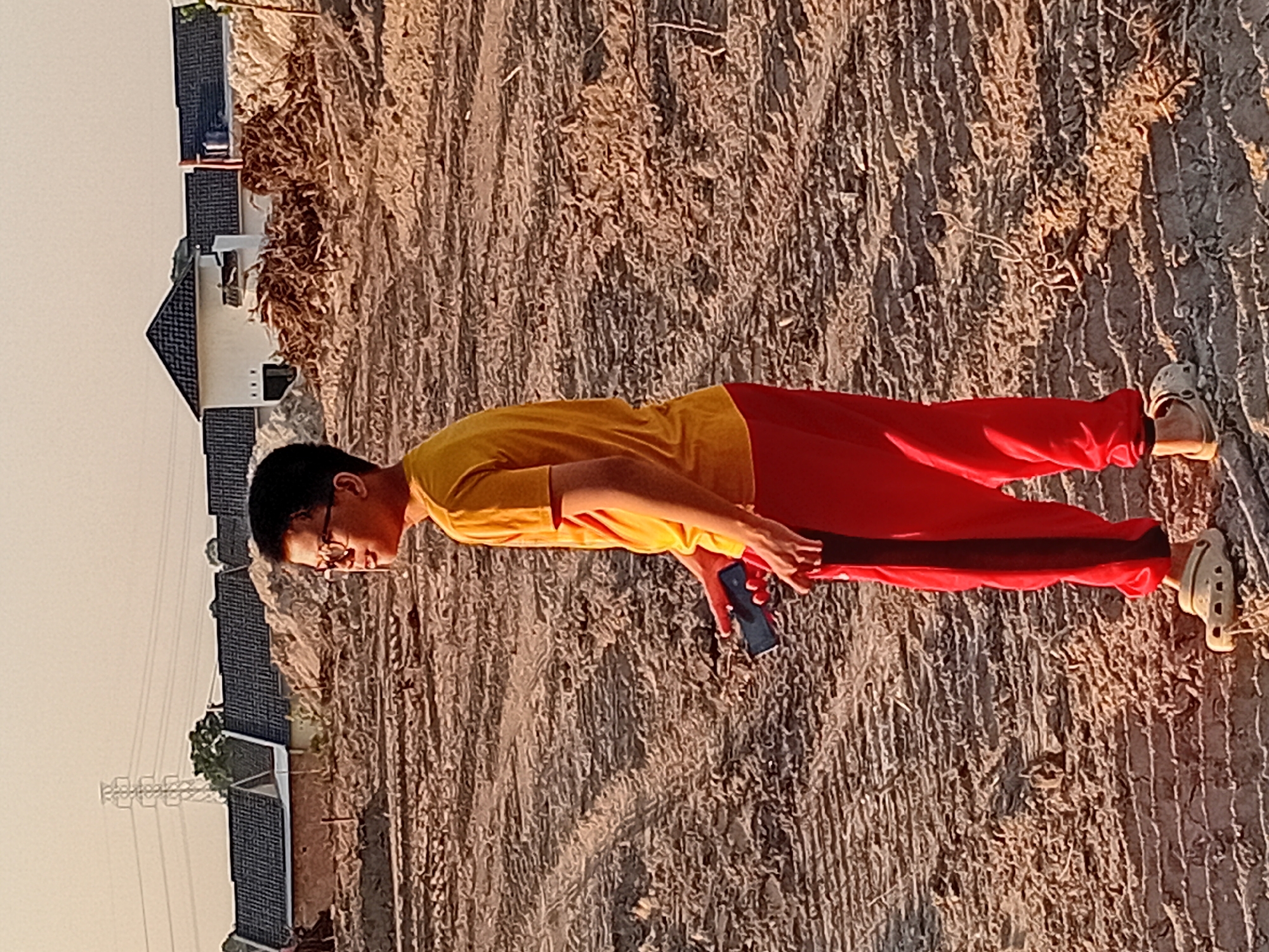 A young person wearing a yellow t-shirt and red pants stands in a dirt field. The person is looking to the right and holding a dark-colored mobile phone in their left hand. The background shows a row of houses under construction with dark tiled roofs, piles of dirt and debris, and a power transmission tower in the distance. The ground is uneven with tire tracks visible. The lighting suggests it is either sunrise or sunset. The image appears to have been taken in Kasang Pudak, Indonesia, given the context.