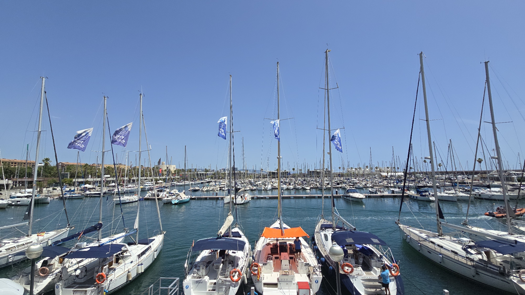 This is a high-angle, wide-shot photograph of a crowded marina on a clear, sunny day, likely in Barcelona, Spain, given the presence of numerous sailboats and the architectural style of the buildings in the background which are reminiscent of Mediterranean coastal cities. The foreground is dominated by several white sailboats docked side-by-side. These boats feature prominent masts, sails furled or covered, and blue canopies over their cockpits. Some of these boats have orange life preservers attached to their railings. One sailboat in the middle foreground has a person in a blue shirt standing on its deck, appearing to be attending to something. Another sailboat to the right has a person in a similar blue shirt visible.
Beyond the foreground boats, the marina extends far into the distance, packed with hundreds of other sailboats and some motorboats. The water is a clear, turquoise blue. In the middle ground, several blue flags with white lettering are attached to the masts of some boats. The background shows a city skyline with low-rise buildings, many with terracotta-colored roofs, and palm trees. The sky is a vibrant, cloudless blue. The overall impression is one of a bustling leisure harbor.
