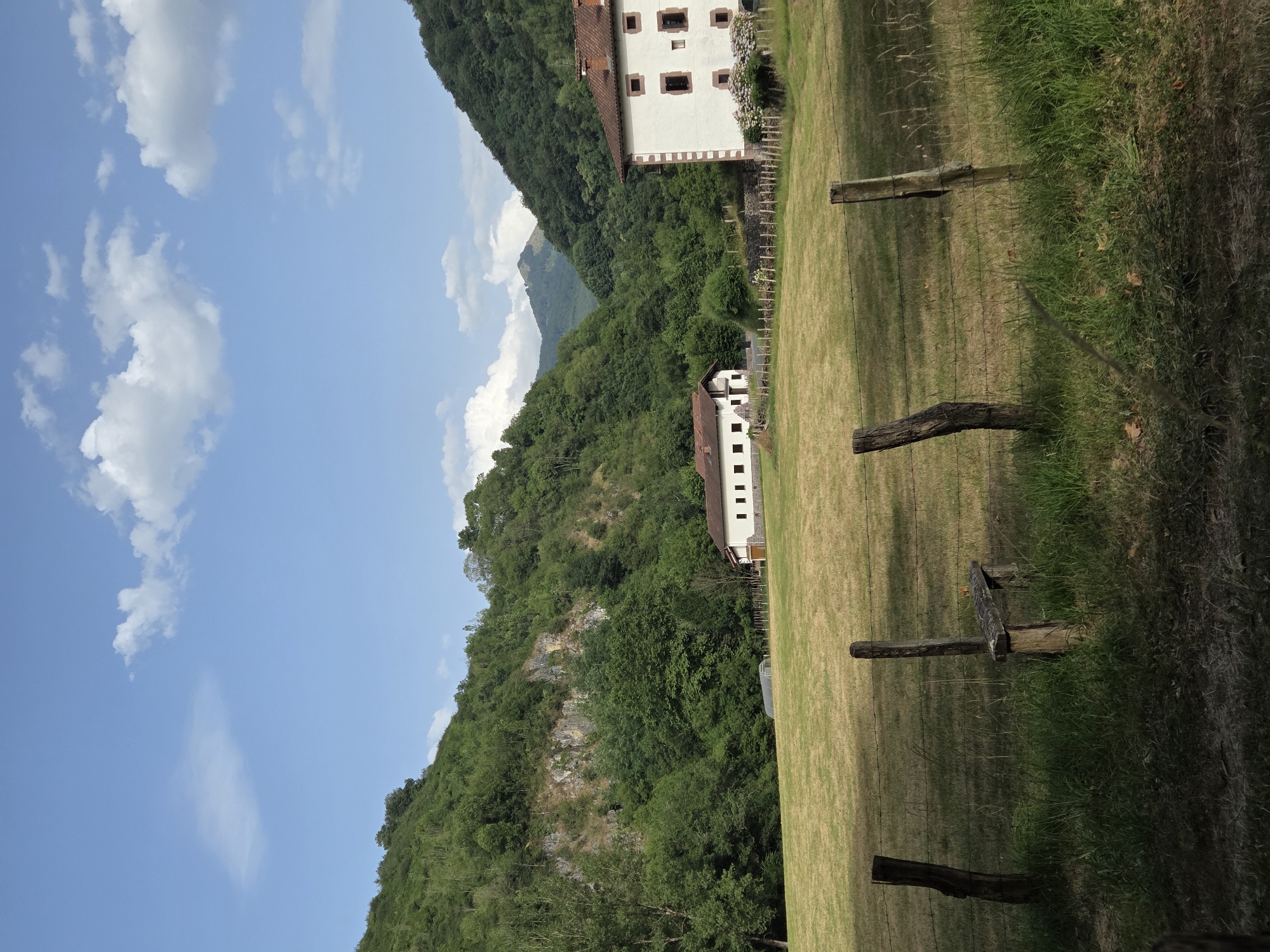 The image depicts a pastoral landscape in the municipality of Baztan, Spain. In the foreground, a rustic wooden fence with several posts and barbed wire runs horizontally across the frame. Beyond the fence, a field of dry, golden grass stretches towards two white buildings with red tile roofs. The building on the right is larger and features a decorative white border around its windows and roofline, with several small, dark windows visible. The building on the left is simpler, with a similar red tile roof and several dark windows.
Behind the buildings, a lush green forest covers the steep slopes of hills that rise dramatically on either side of a valley. A rocky outcrop is visible on the left-hand hillside. In the distance, the valley opens up to reveal a hazy mountain peak under a partly cloudy blue sky. A small greenhouse structure is partially visible in the field between the fence and the buildings. The scene is illuminated by natural daylight, suggesting it was taken during the daytime. No people are present in the image.