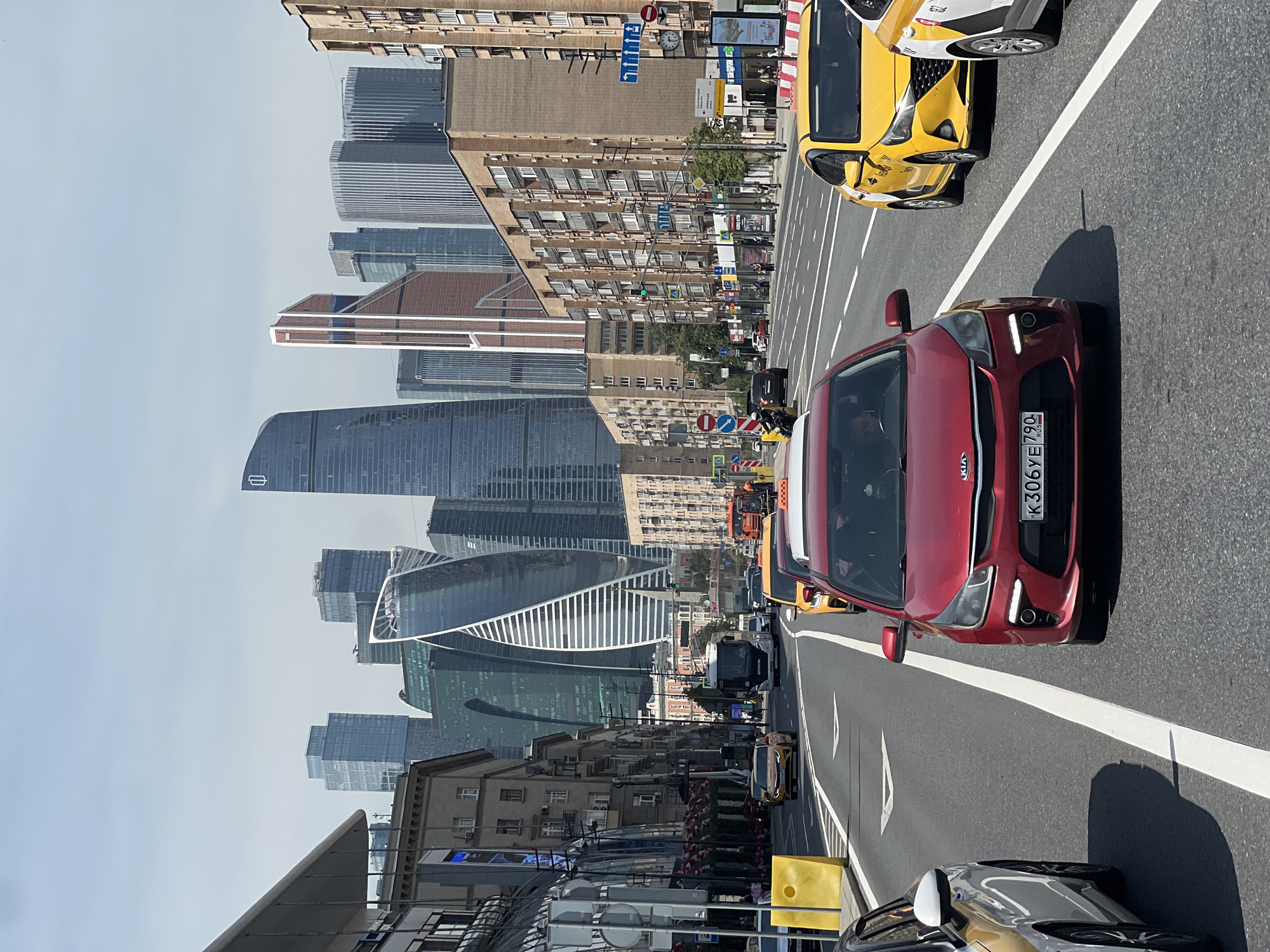 The image depicts a multi-lane urban thoroughfare in Moscow, Russia, viewed from a slightly elevated perspective looking west towards the Moscow International Business Center (Moscow-City). The scene is bathed in bright daylight under a clear, pale blue sky with minimal cloud cover, suggesting fair weather.
In the immediate foreground, a dark red Kia Rio (license plate K306YE 790 RUS) occupies the center lane, heading away from the viewer. Two occupants are visible inside the vehicle. To its right, a yellow Hyundai Solaris taxi follows, and partially visible to the far right is a white vehicle with yellow and black markings, likely another taxi. On the far left, the front quarter of a light-colored car is visible. The asphalt road features clear white lane markings and a double solid white line separating the main lanes from what appears to be a dedicated bus or taxi lane on the left. Yellow concrete barriers are present along the left edge of the road, bordering a sidewalk.
Further down the road, a white Scania bus with "SCANIA" text on its front is visible in the left lanes, followed by other vehicles, including several yellow taxis. On both sides of the street, multi-story buildings line the thoroughfare. The buildings on the right are primarily light brown brick, with numerous windows and balconies, consistent with residential or mixed-use structures. On the left, a modern building with a curved, reflective glass facade stands alongside more traditional, older Moscow architecture. Street furniture, including lampposts, traffic lights (one green light is visible on the right), and various street signs are present. A blue directional sign with upward arrows and a red circular "no entry" sign are discernible on the right side. Pedestrians can be seen on the sidewalks.
Dominating the distant background is the distinctive skyline of the Moscow International Business Center. Prominent skyscrapers include the tall, dark, cylindrical OKO Tower North with a white logo near its summit, the spiral-shaped Evolution Tower, and the geometric, multi-faceted Mercury City Tower with its golden-bronze hue. Various other modern glass and steel high-rises complete the imposing cluster of buildings.
The presence of the Moscow-City skyline, Moscow region license plates, and typical Moscow street architecture indicates the location is along Kutuzovsky Prospekt, looking west.