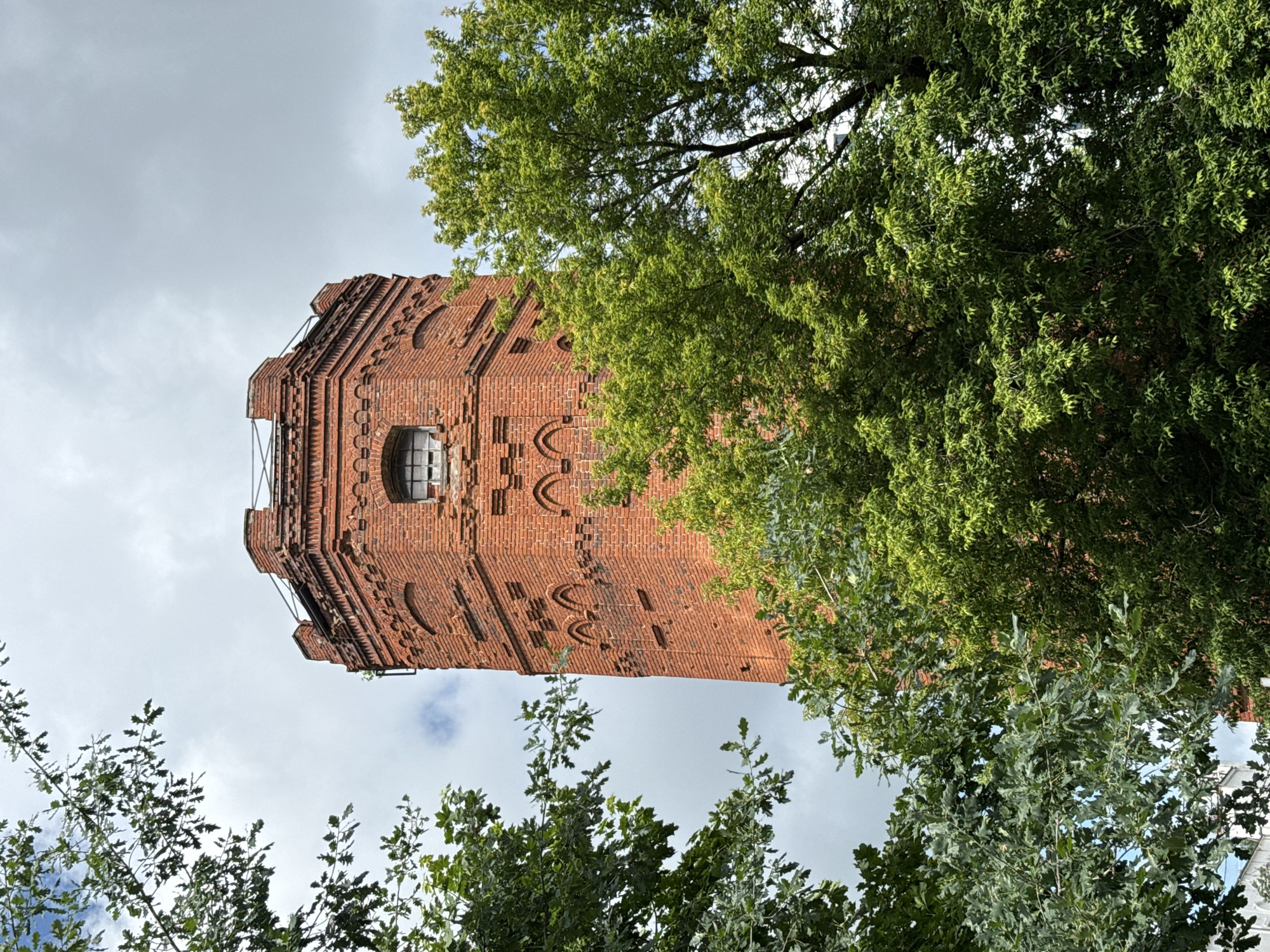 The image shows the top portion of a red brick water tower, an octagonal structure with a crenellated top, framed by lush green trees and a cloudy sky. The tower features decorative brickwork, including arched elements, and a small, square window with a dark frame. A thin metal railing runs along the perimeter of the tower's top. The trees, with dense foliage, partially obscure the tower, creating a natural framing effect. The overall impression is of an old industrial structure integrated into a verdant, possibly park-like, setting. This water tower is located in Izhevsk, Russia.