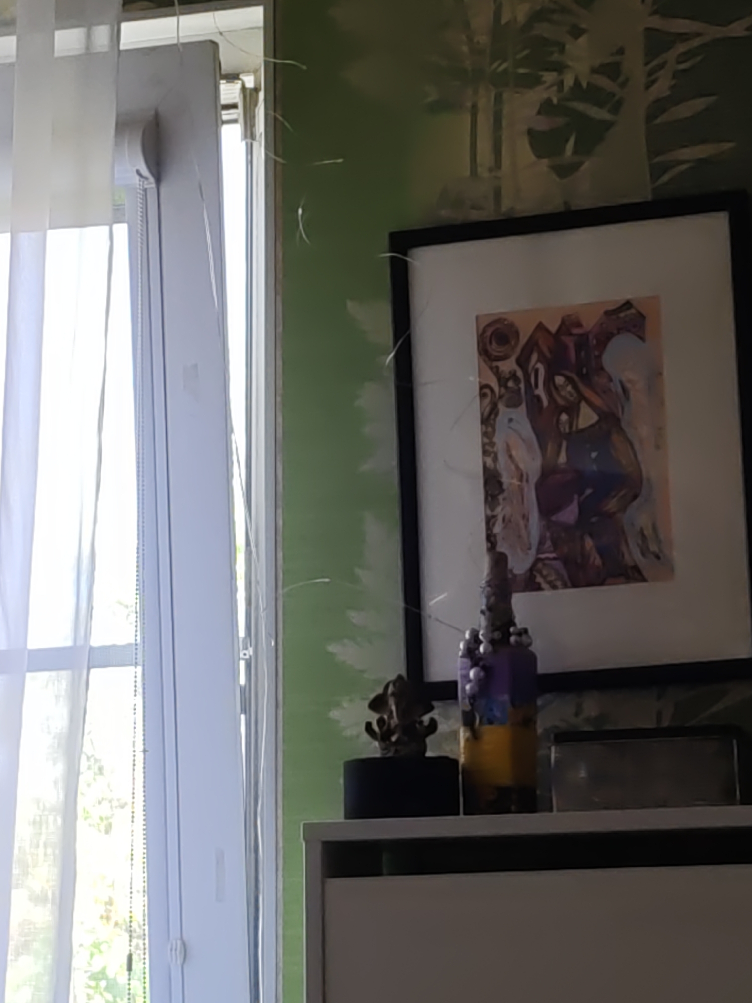 This is a medium shot of an indoor scene, likely a room, taken with a handheld camera, resulting in a slightly blurred and grainy image. The focus is on a shelf against a green wall with a white tree pattern. On the shelf, from left to right, is a small, dark, ornate figurine on a black base, a tall decorative bottle with purple and yellow sections adorned with small white beads, and a rectangular dark object, possibly a picture frame or a box. Behind the shelf, a framed piece of abstract art with warm tones and dark figures is visible. To the left of the scene, a white window with sheer white curtains is present, with light filtering through. The image quality is poor, making precise identification of all details difficult. The image does not contain any information that can be used to determine its location in Rostov-on-Don, Russia.