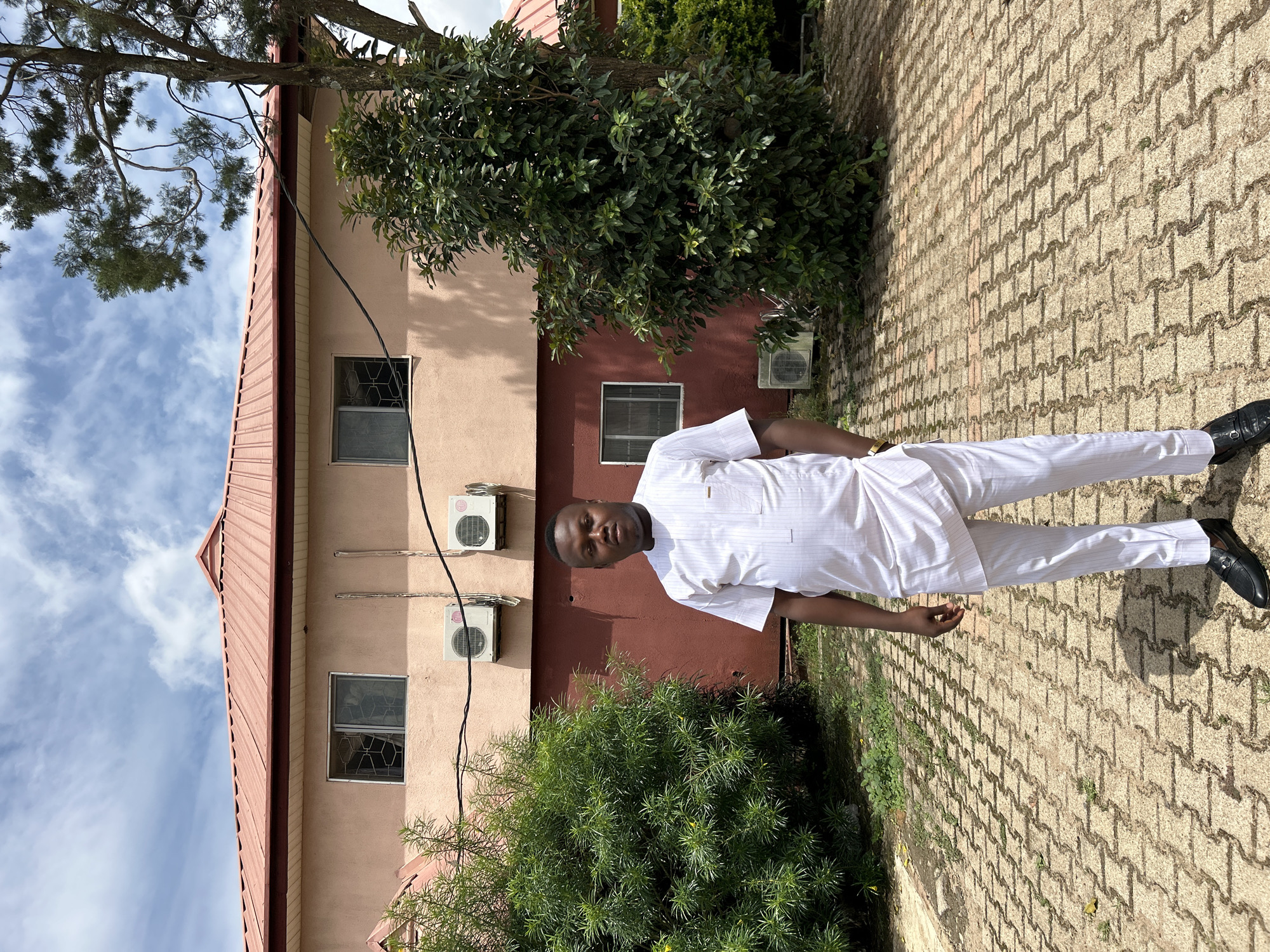 An outdoor, eye-level full-shot depicts an adult male standing centrally in front of a two-story building under a partly cloudy sky in what is identified as Jos, Nigeria. The man, with a medium complexion and short dark hair, wears a white, vertically striped traditional Nigerian tunic and matching trousers, along with black slip-on shoes featuring a metallic embellishment; his hands are in his pockets, and his expression is neutral. The building behind him features a light peach upper facade and a dark red lower facade, topped with a red corrugated metal roof. Security grilles are visible on the windows of both floors. Multiple white split-unit air conditioner outdoor units are mounted on the building's exterior. Lush green shrubs flank the building on both sides, with a larger tree with prominent branches on the right. The foreground is paved with light-colored interlocking concrete blocks.