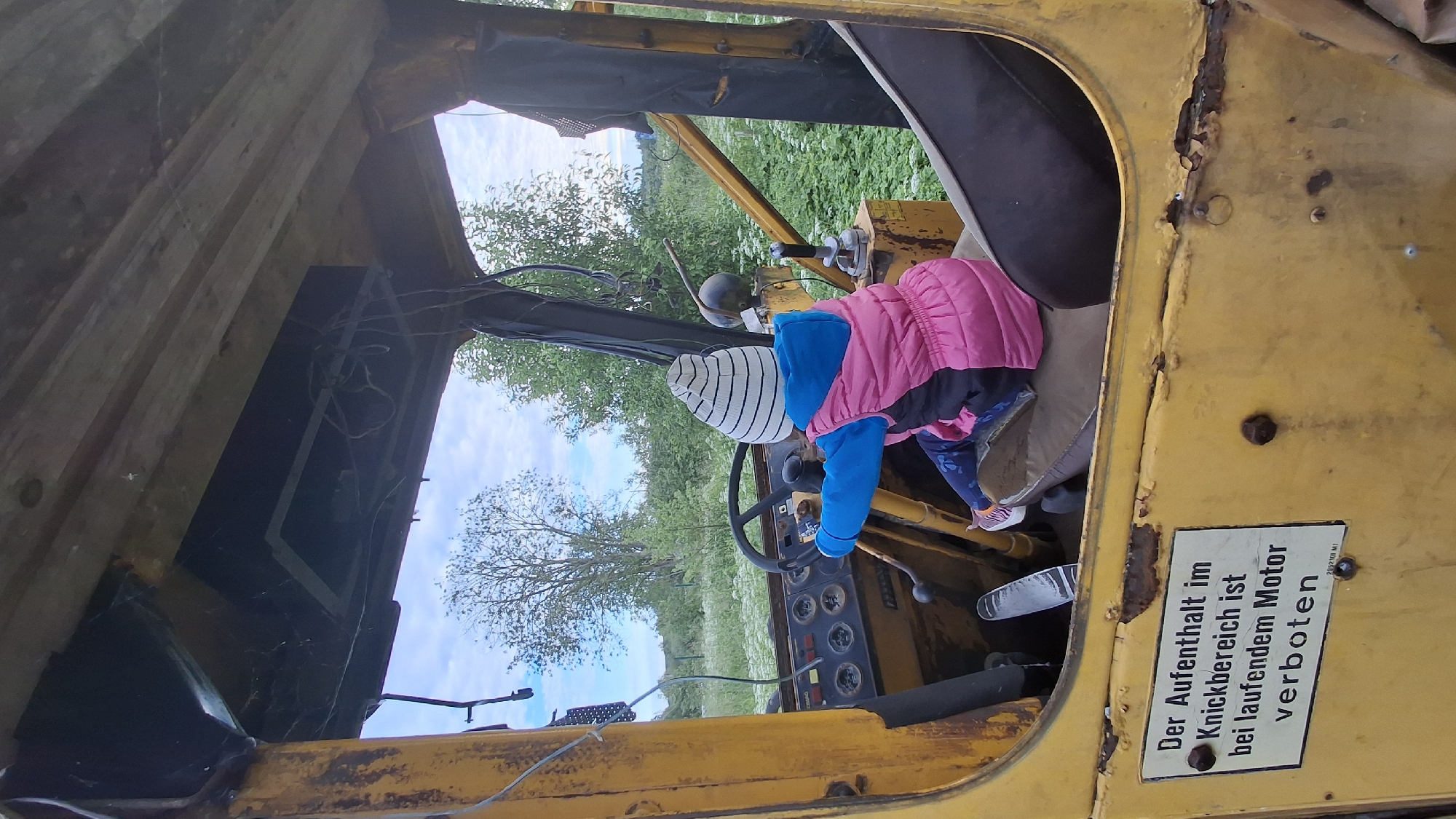 The image shows a young child, wearing a striped beanie, a blue long-sleeved shirt, and a pink puffer vest, sitting in the driver's seat of a weathered, yellow vehicle, possibly an old tractor or construction machine. The child's hands are on the steering wheel. The interior of the vehicle is visible, featuring a dashboard with various gauges, a gear shift, and pedals. A sign with German text, "Der Aufenthalt im Knickbereich ist bei laufendem Motor verboten," is affixed to the exterior yellow panel below the driver's side. The vehicle appears to be outdoors, with a background of green foliage and a cloudy sky. The location is identified as Innolovo, Russia.