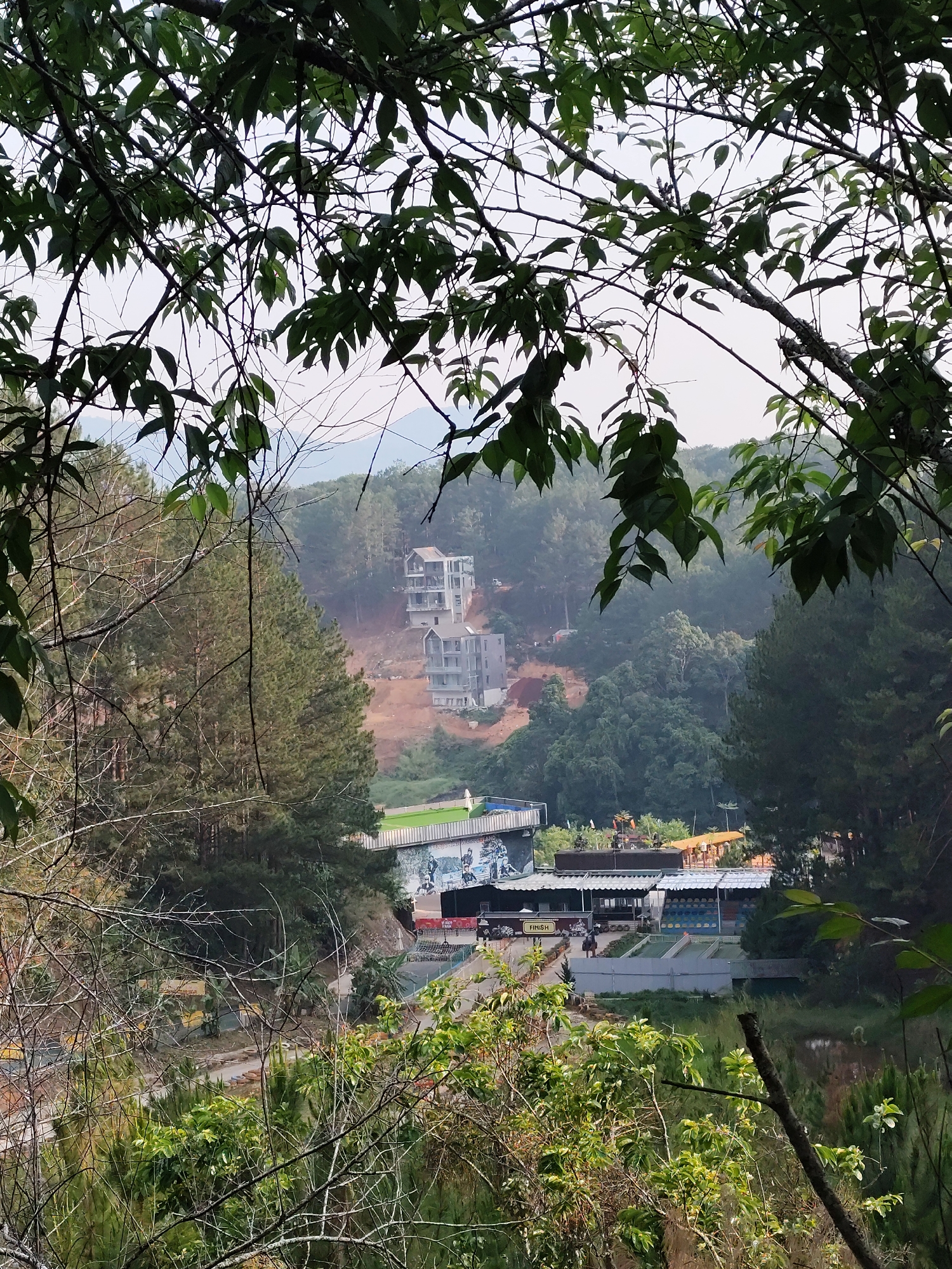 The image depicts a view from a vantage point in Da Lat, Vietnam, looking down at a developed area surrounded by lush green hills. The foreground is filled with tree branches and foliage, framing the scene. Below, there's a complex of buildings, structures, and landscape features. A multi-story building is situated higher on the hill, appearing to be under construction or recently built. Lower down, the complex has multiple structures with what looks like a covered outdoor seating area, possibly a restaurant or recreational facility. The scene is set in a mountainous region. The area appears to be a tourist spot with recreational facilities. There are no people prominently visible. The environment is misty, suggesting a humid, possibly high-altitude location.