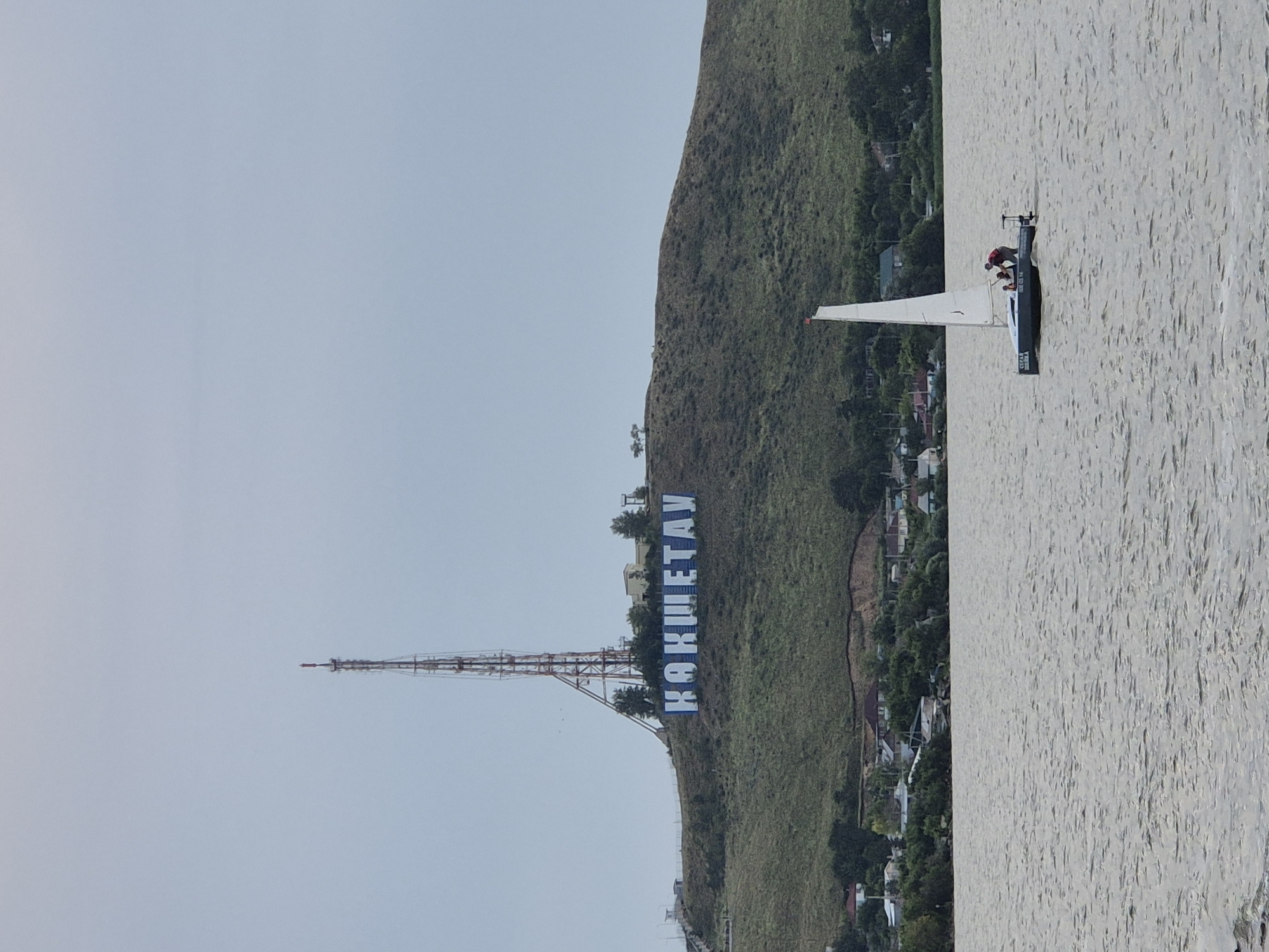 An aerial view of the city of Kokshetau, Kazakhstan, shows a wide body of water with a sailboat in the foreground. The sailboat has a white sail and a gray hull, with two people on board. The water is rippling, indicating a slight breeze. In the background, a hill is visible, with large blue letters spelling out "KOKSHETAV" on its side. A tall communication tower stands at the crest of the hill. Several houses and trees are scattered along the base of the hill and on the shoreline. The sky is overcast, creating a muted light over the scene.