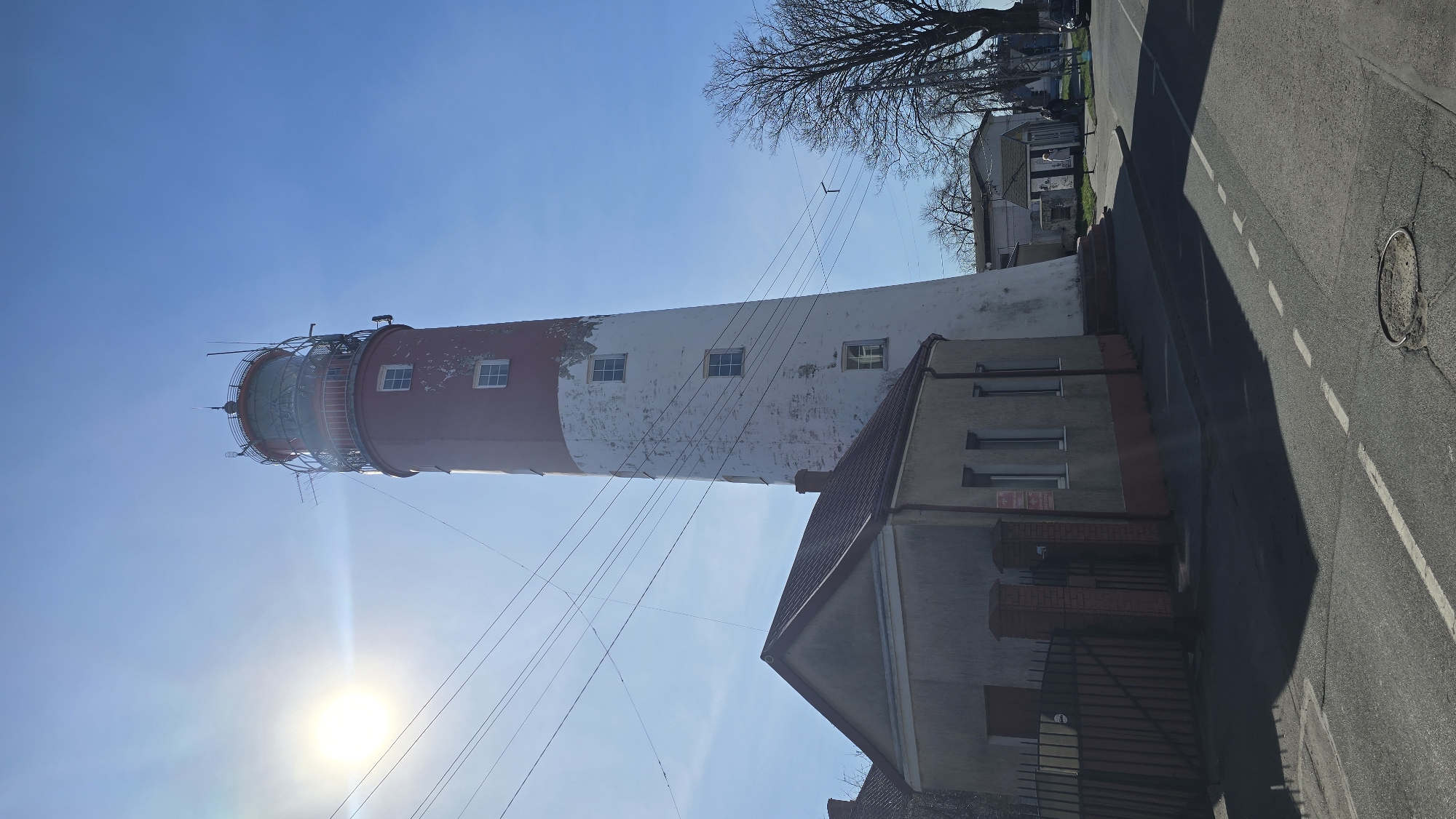 The image shows the Baltiysk Lighthouse, located in Baltiysk, Russia. The lighthouse is painted with alternating bands of white and red. It is a tall, cylindrical structure with a visible lantern room at the top, surrounded by a metal cage. A few windows are visible along the body of the lighthouse. The lighthouse is situated next to a smaller, gray building with a red roof and a fenced area. Power lines stretch from the lighthouse towards the foreground. The sky is clear blue with the sun shining brightly. A paved road runs along the right side of the image. Trees and other buildings are visible in the background.