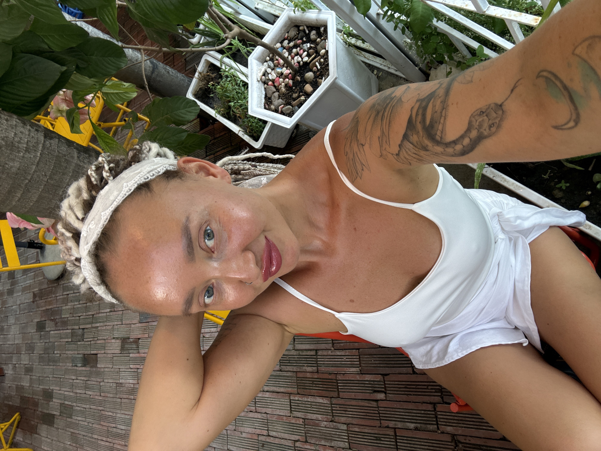 A woman is taking a selfie, resting her head on her hand. She has blonde dreadlocks and a white headband. She is wearing a white tank top and white shorts. There is a tattoo of a snake on her left arm. She is sitting outside, with a brick patio behind her and some plants and planters. The scene appears to be outdoors, possibly in a garden or patio area. The lighting suggests it is daytime. The woman's expression is relaxed and casual. There is no visible text in the image.