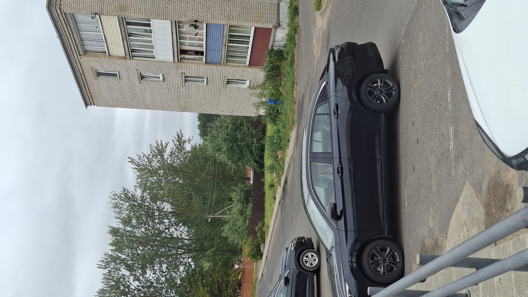 A black sedan is parked on a paved surface in front of a multi-story brick apartment building. The building has several windows and balconies. In the background, there are trees and a cloudy sky. Another black car is partially visible in the foreground to the left, and the hood of a white vehicle is in the lower right corner. A metal railing is in the immediate foreground. This scene is located in the city of Smolevichi, Belarus.