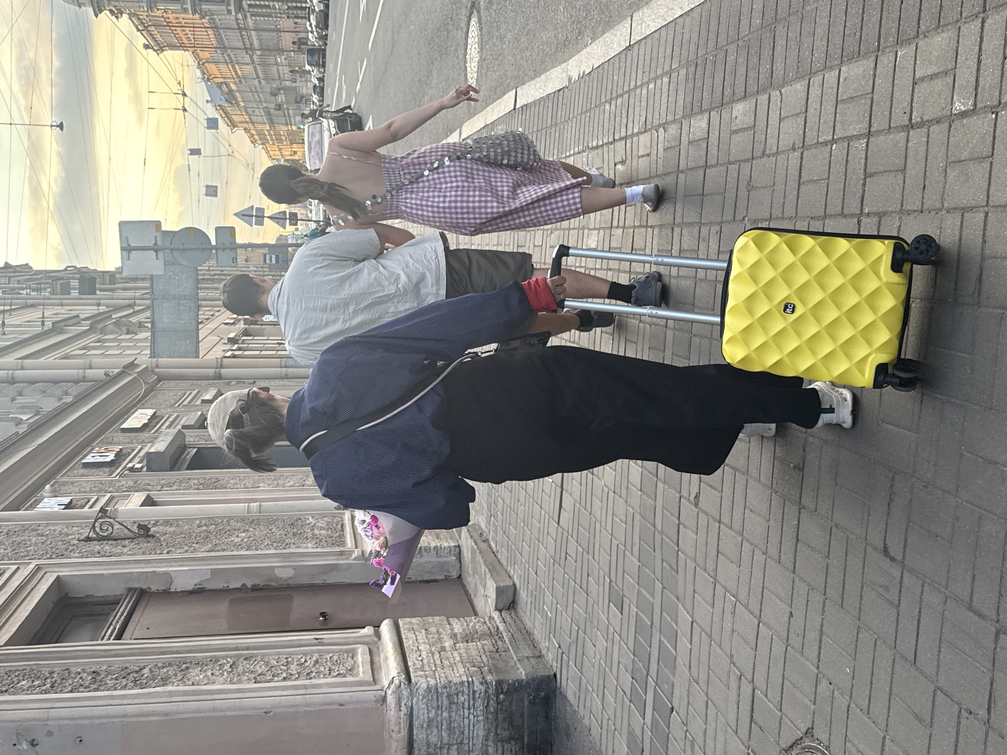The image shows three people walking on a cobblestone sidewalk in Saint Petersburg, Russia. A woman in a pink and white checkered dress and a sequined bag is walking ahead, with her right hand raised in a peace sign. She is followed by a man in a light-colored shirt and dark shorts. Behind them, another person, dressed in dark clothing and a blue and white striped shirt, is walking and pulling a bright yellow suitcase with a textured pattern. This person is also holding a bouquet of flowers.
The scene appears to be set in the late afternoon or early evening, as indicated by the warm, soft light illuminating the buildings and the sky, which is a pale yellow and orange. The architecture of the buildings suggests an urban environment, with classical detailing and multiple stories. Overhead wires and streetlights are visible, hinting at a bustling city. The weather seems pleasant, with no signs of rain or strong winds. The overall mood is one of casual movement through the city, possibly on a journey or heading to an event. No visible text is discernible in the image.