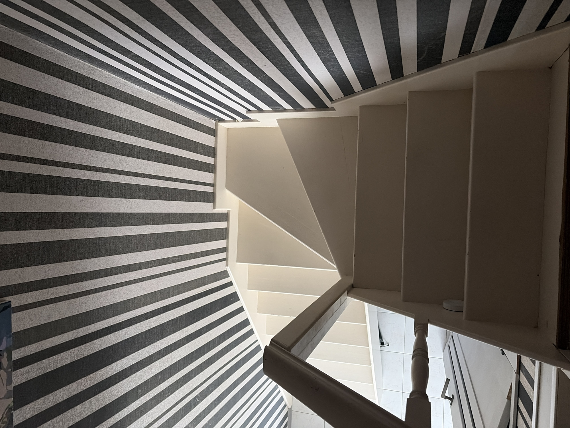 The image shows a staircase with white steps and a white banister, ascending into the frame. The walls and ceiling are decorated with black and white striped wallpaper. There are no people visible in the image. The lighting suggests an indoor environment, likely a residential building. The specific location in Sochi, Russia, cannot be determined from the provided image.