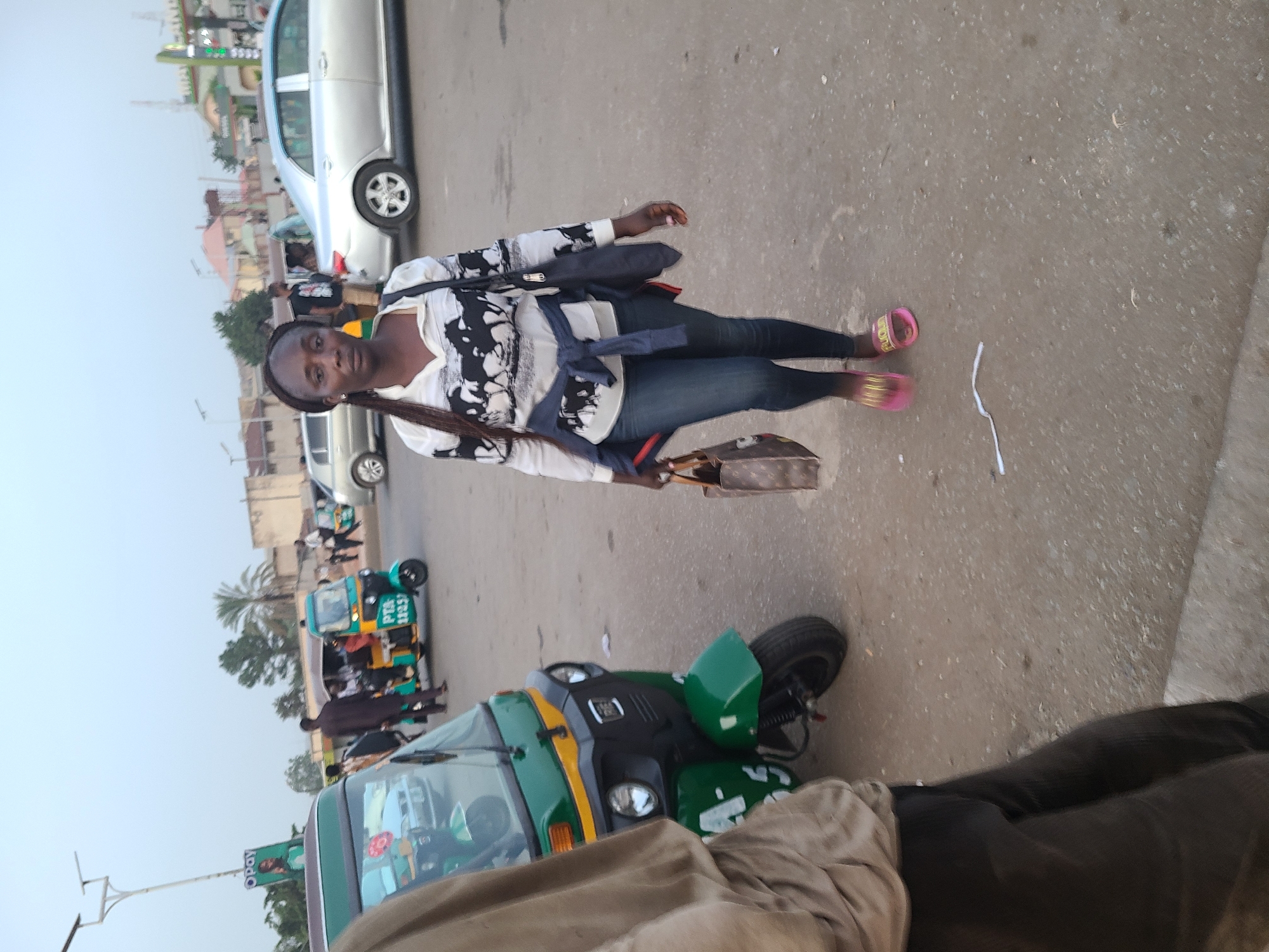 A woman is standing in the street in Jos, Nigeria. She is wearing a black and white patterned shirt, blue jeans, and pink sandals. She is carrying a brown handbag and a black satchel. A green and yellow tricycle is parked nearby. In the background, there are other vehicles and buildings.