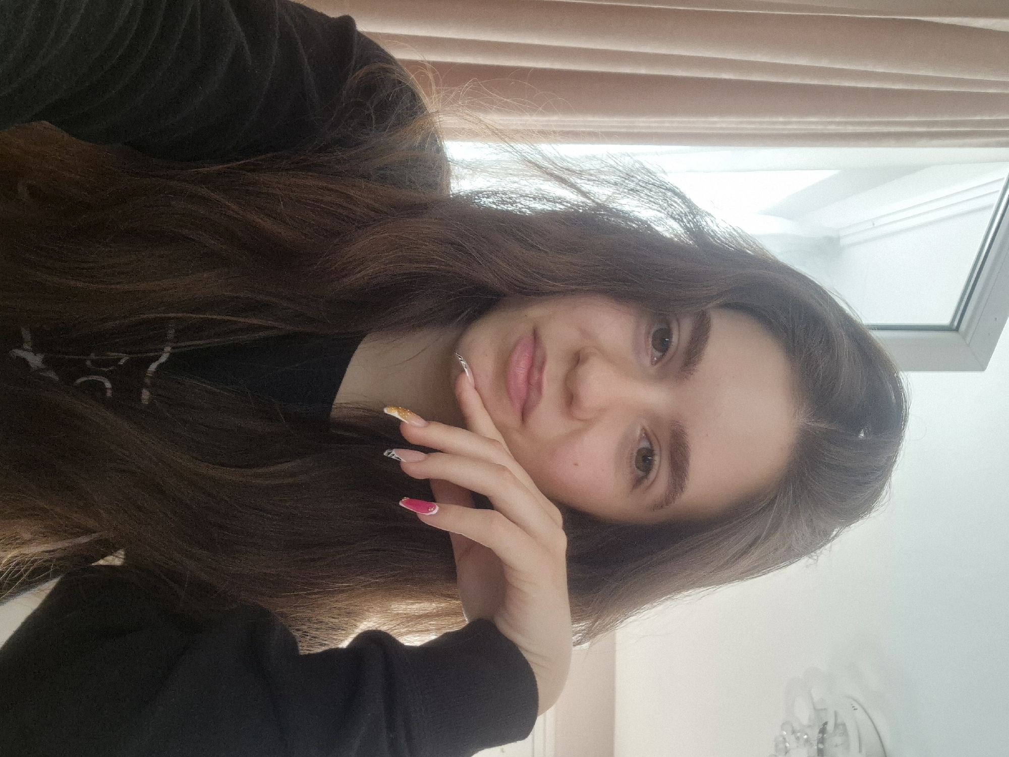 A young woman is posing for a selfie. She has long, brown hair and is wearing a black sweater. Her hand is resting on her cheek. She has manicured nails with different designs. The background includes a window with closed blinds and a white wall. The location cannot be determined from the image.