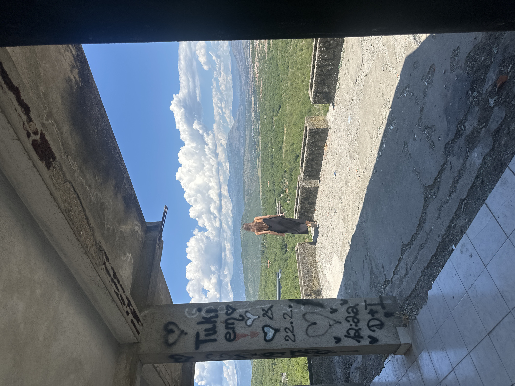 A woman in a black dress stands on a concrete balcony overlooking a valley in Gornji Kokoti, Montenegro. The balcony is part of an unfinished concrete structure, with graffiti visible on a supporting pillar. The woman is seen from behind, walking away from the camera towards the scenic landscape. The background features rolling green hills, mountains, and a bright blue sky with white clouds.