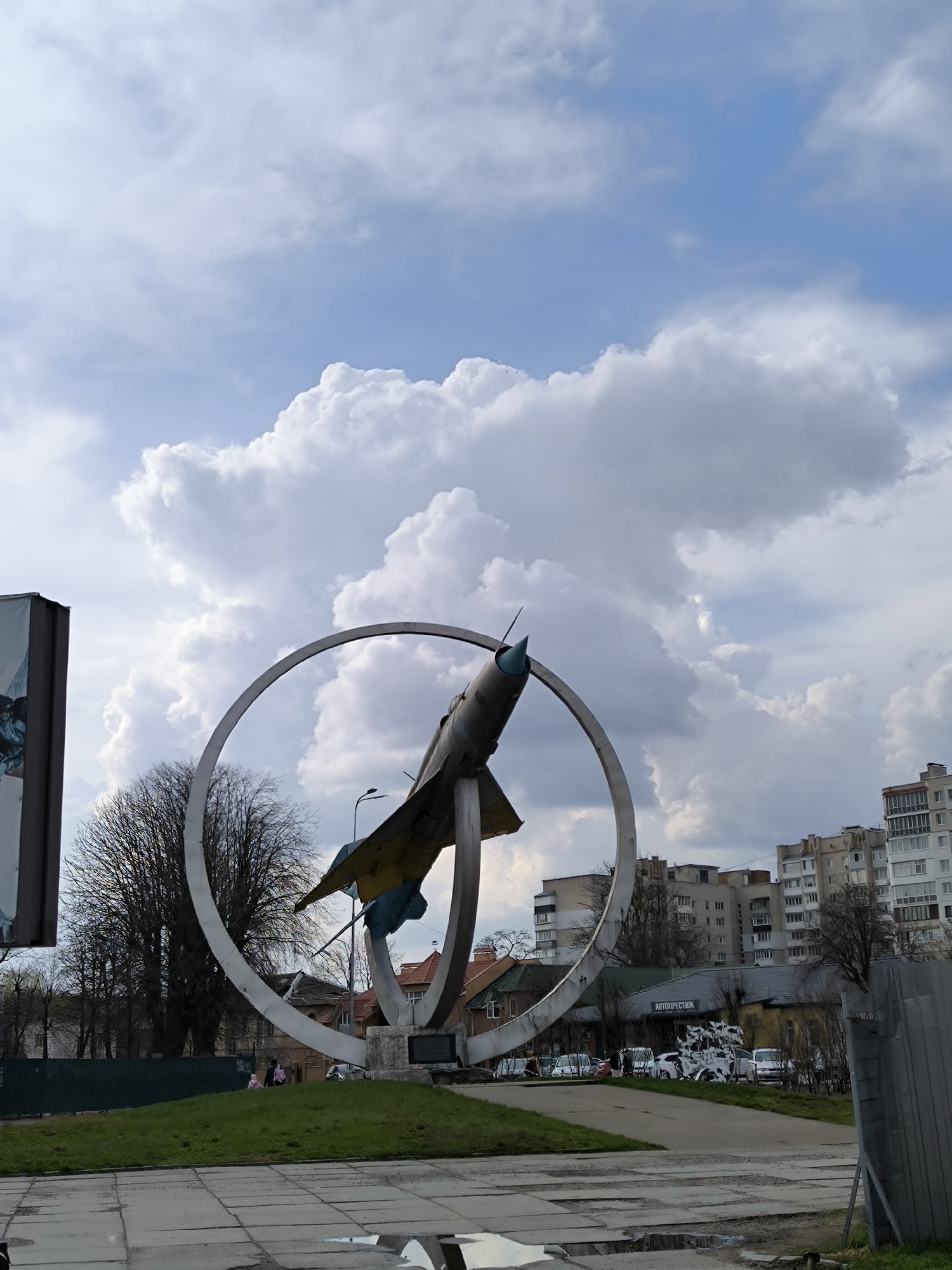 The image shows a Soviet MiG-21 jet fighter mounted as a monument on a pedestal within a circular metal ring. The jet is painted in a yellow and blue camouflage pattern, reflecting the colors of the Ukrainian flag. The monument is located in Vinnytsia, Ukraine, near a road junction. Buildings of varying heights surround the area, including several apartment blocks. Bare trees are visible. The sky is filled with large, puffy white clouds against a blue backdrop. The ground appears to be paved with gray concrete.