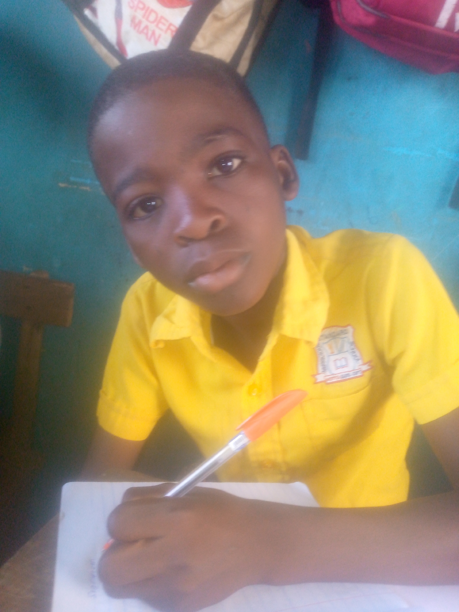 A young boy with dark skin and short hair is sitting at a desk, writing in a notebook with a pen. He is wearing a bright yellow collared shirt with a school emblem on the pocket. The background includes a blue wall, a "Spider-Man" bag, and a red bag hanging behind him. The location is Unknown, Unknown.