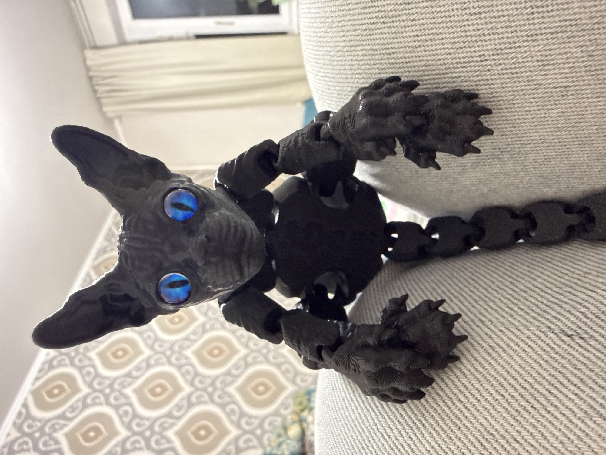 A black, articulated toy resembling a hairless cat is perched on a surface covered in light-grey fabric. The toy cat has large, blue eyes and a segmented body. The surface appears to be a piece of furniture. The background features a wall with patterned wallpaper and a window with closed blinds. It is not possible to determine the location as Khimki, Russia from the available information.