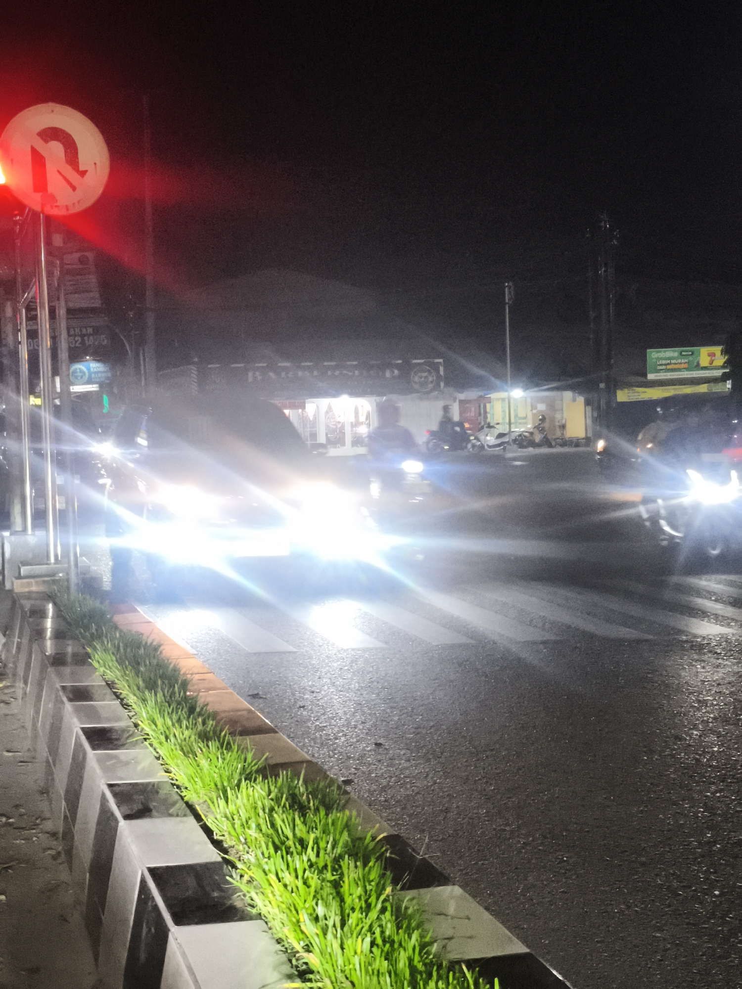 This is a night-time, medium-shot, eye-level photograph taken on a wet road in Samarinda, Indonesia, likely at an intersection with a pedestrian crossing. The scene is illuminated by streetlights and the headlights of passing vehicles. In the foreground, to the left, a traffic light is visible, with the red light illuminated and a "U-turn prohibited" sign next to it. A decorative planter with bright green grass runs along the edge of the road. The road surface is dark and wet, reflecting the lights. A pedestrian crossing is marked with white stripes across the road. In the midground, several motorcycles and at least one car are on the road, their headlights creating bright streaks of light. Some buildings are visible in the background, with lights on, suggesting they are shops or businesses. One building appears to have a sign that says "BARBERSHOP". Another sign, possibly an advertisement for a ride-sharing service, is visible in the far background on the right. The overall atmosphere is that of a busy urban street at night, with the rain adding a slick sheen to the pavement.