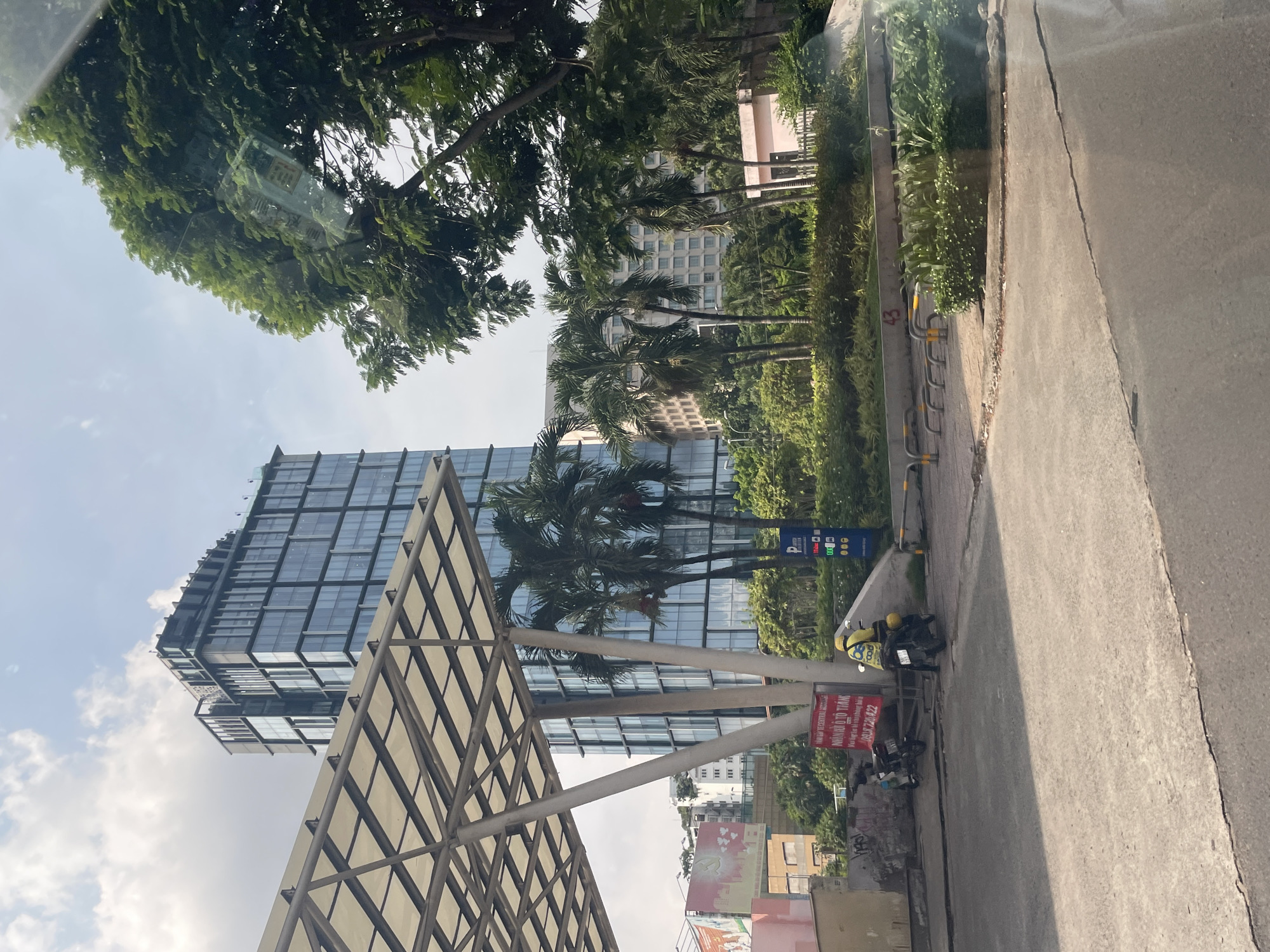 The image captures a street scene in Thủ Đức, Vietnam, on a partially cloudy day. In the foreground, a gray asphalt road shows cracks and wear. To the left, a covered structure with a white canopy supported by angled beams extends into the frame. Beyond this, a tall, modern glass-facade building dominates the background, with its reflective windows displaying the sky. In front of this building, palm trees and lush green vegetation form a natural buffer.
To the left of the covered structure, a pink and beige building is visible, with some graffiti on its lower wall. A red and white sign with Vietnamese text and a phone number is mounted on a concrete barrier. Two motorbikes are parked near this barrier; one is black and the other is a lighter color.
In the mid-ground, a person wearing a yellow jacket and helmet is riding a black motorbike. This motorbike has a yellow delivery box on the back with the word "FOOD" in black letters. To the right of this motorbike, a blue vertical sign with white text and graphics is visible, possibly indicating parking or fuel prices. Further to the right, more trees and greenery are present, with a low pink building peeking through. The scene suggests a commercial or urban environment with elements of both modern architecture and tropical vegetation.