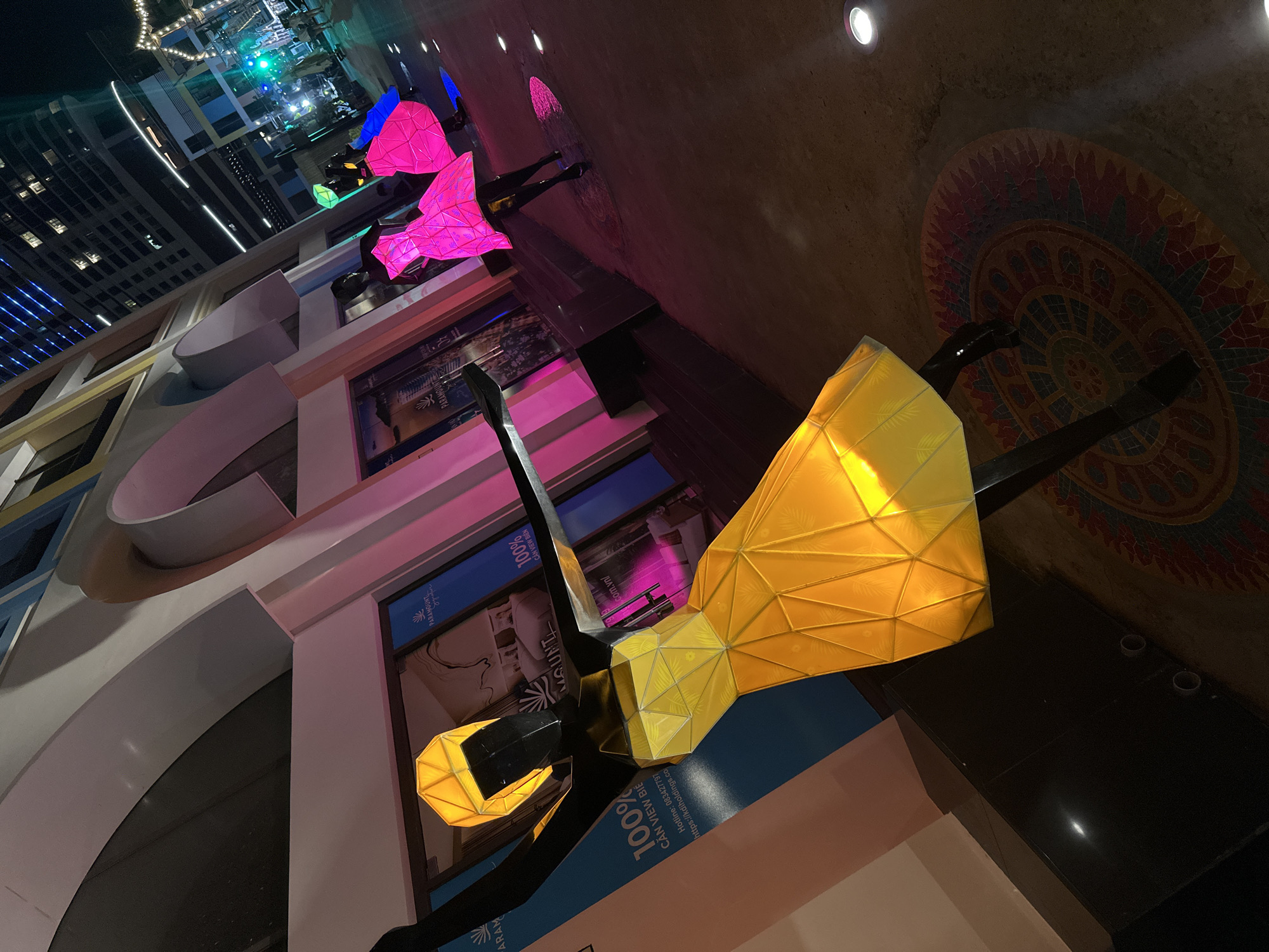 The image captures a night scene in Bac Nha Trang Ward, Vietnam, featuring architectural details and decorative illuminated sculptures. Dominating the foreground is a bright yellow, geometrically-shaped sculpture of a person, situated in front of a building with advertisements that read "PARAMOUNT 100% CAN VIEW BIẾN" with a website and hotline. Additional sculptures, illuminated in varying shades of pink and blue, are visible further in the background. The building's architecture includes rounded arches and shop windows. The ground surface has a decorative circular design, and overhead lights illuminate the area. In the background, taller buildings with illuminated windows can be seen.