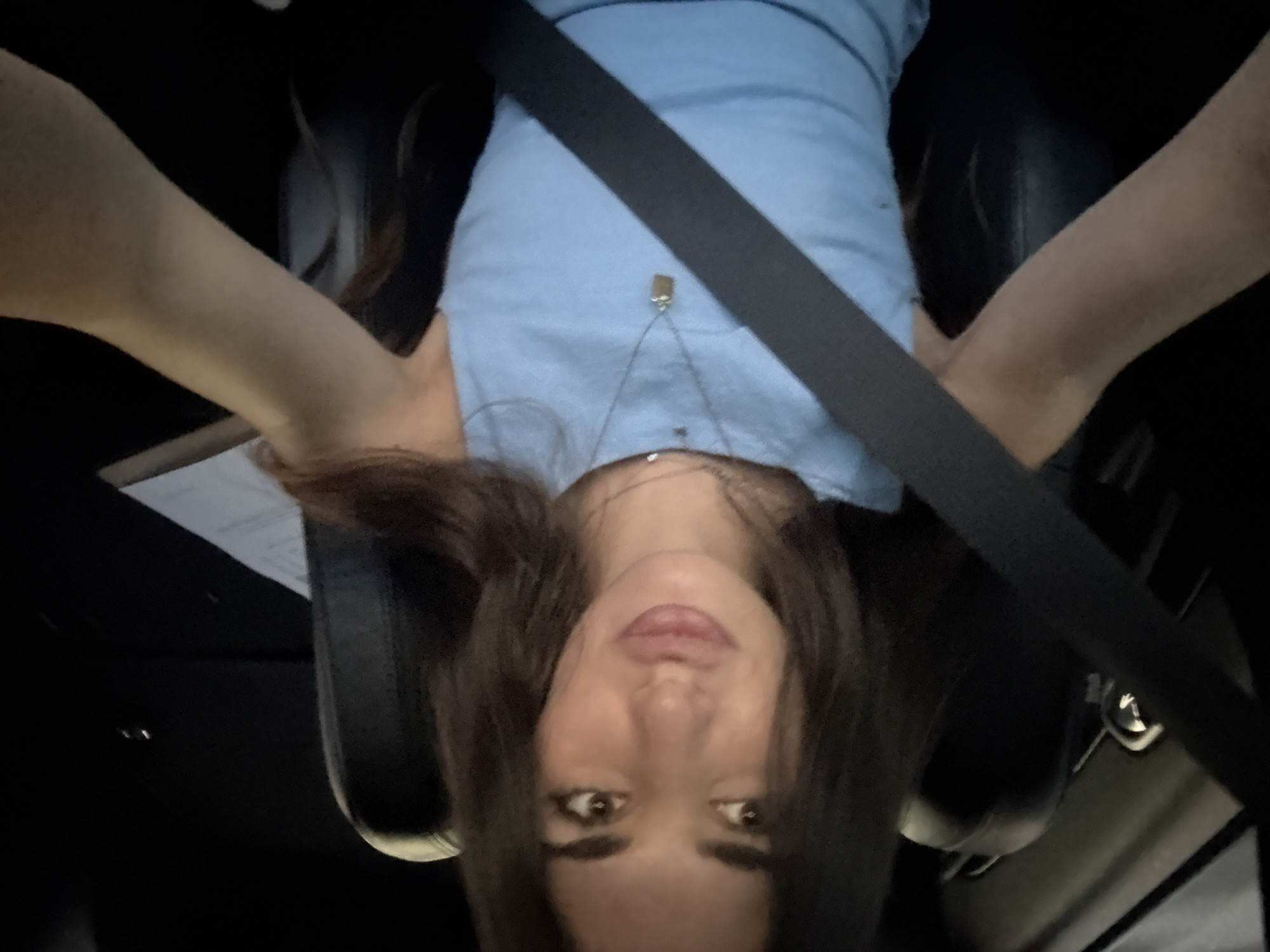 A young woman with long, dark hair is depicted in a selfie taken inside a vehicle in Boca Raton, United States of America. She is seated, wearing a light blue sleeveless top and a dark seatbelt diagonally across her chest and shoulder. Her lips are slightly puckered, and she looks directly at the camera. She wears two necklaces, one choker-style and another with a rectangular, metallic pendant. The vehicle interior features dark leather seats, and the overall environment appears dimly lit, possibly at night. A white document or notepad is visible on the passenger seat to her right. Her arms are extended, holding the camera to capture the image.