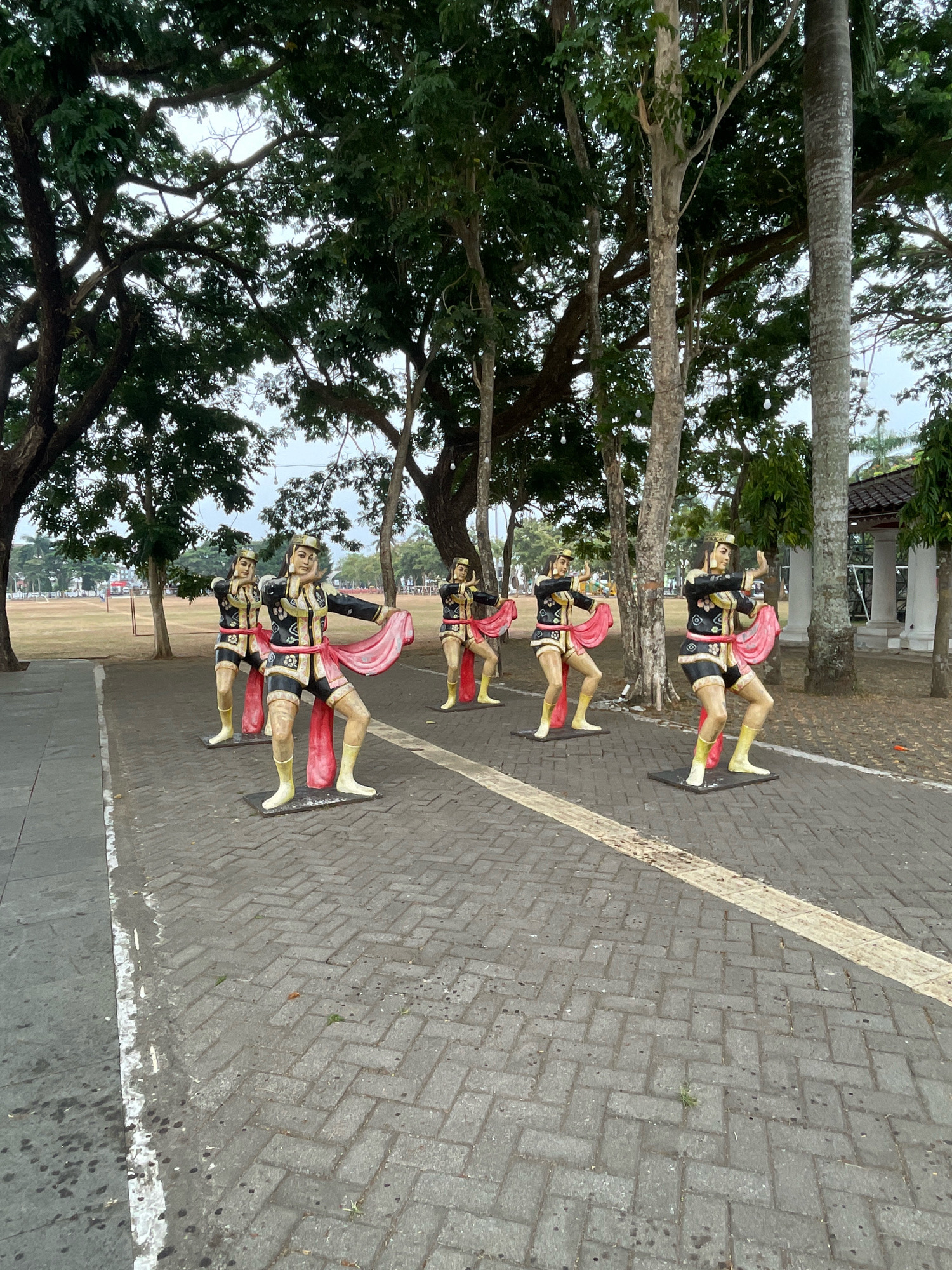 This image depicts a row of six life-sized statues of dancers posed in a traditional Indonesian dance. The statues are positioned on a paved walkway made of interlocking gray bricks, with a white stripe marking the edge of the walkway. The dancers are dressed in ornate black and gold costumes with red sashes and yellow footwear. They are arranged in a staggered formation, facing slightly towards the viewer.
Behind the statues, large trees with dense green foliage provide a canopy. The ground beyond the walkway is a large, open grassy field. In the background, a building with white columns and a dark roof is visible, suggesting a public space or park. The sky is overcast.
This scene is located in the city of Purworejo, Indonesia, specifically at Alun-Alun Purworejo, a central town square often adorned with cultural art installations.