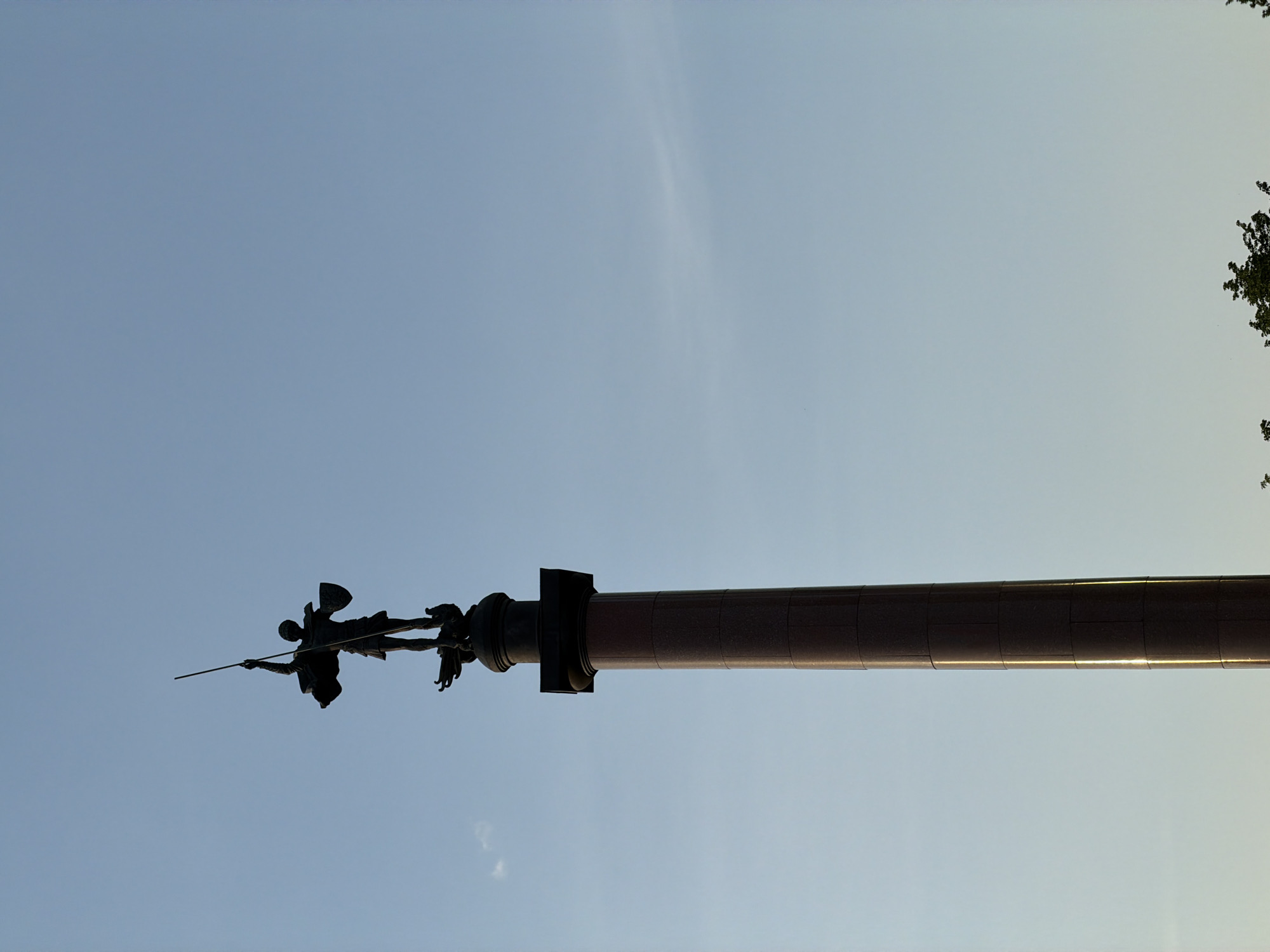 A ground-level, upward-facing shot depicts the Victory Monument in Moscow, Russia. The monument features a tall, cylindrical column, likely made of a dark reddish-brown stone, topped with a statue of Saint George slaying a dragon. The statue and the upper portion of the column are silhouetted against a light blue sky. The angle of the shot makes the monument appear horizontal. A few tree branches are visible in the upper right corner of the image.