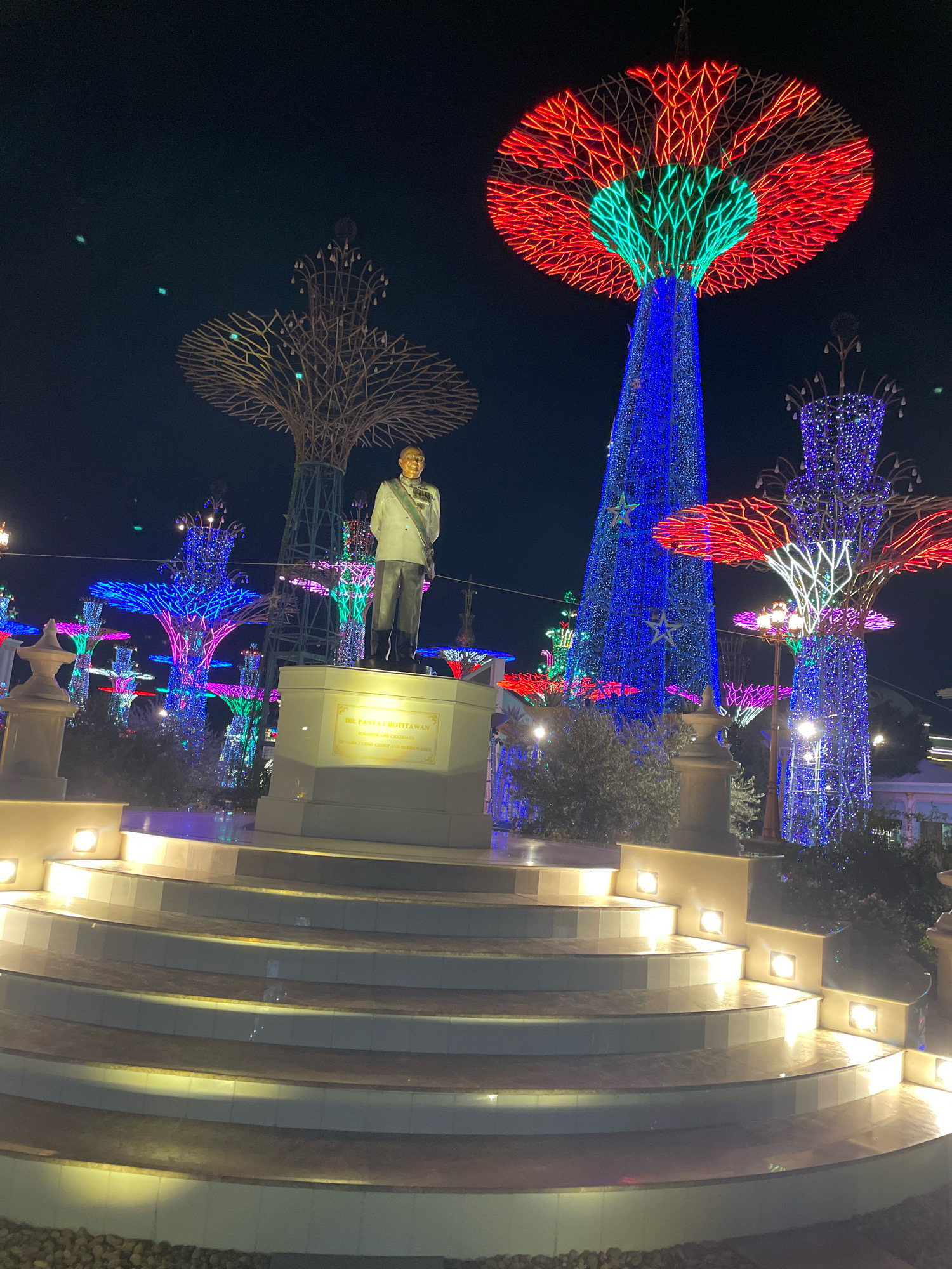 The image depicts a nighttime outdoor scene in Pattaya City, Thailand, featuring a statue of a man in a formal suit and sash, identified by a plaque as "DR. PANYA HUTTAWAN PRESIDENT AND CHAIRMAN". The statue stands on a tiered, illuminated platform with curved steps. Behind the statue, the landscape is dominated by illuminated, tree-like structures adorned with numerous colorful lights, primarily in shades of blue, red, and green. One prominent tree-like structure behind the statue is a tall, cone-shaped display covered in blue lights with stars, topped with a large, circular canopy of red and green lights. Other similar, but smaller, illuminated tree structures are visible throughout the scene, creating a festive and vibrant atmosphere. The ground around the base of the steps is covered with small stones.