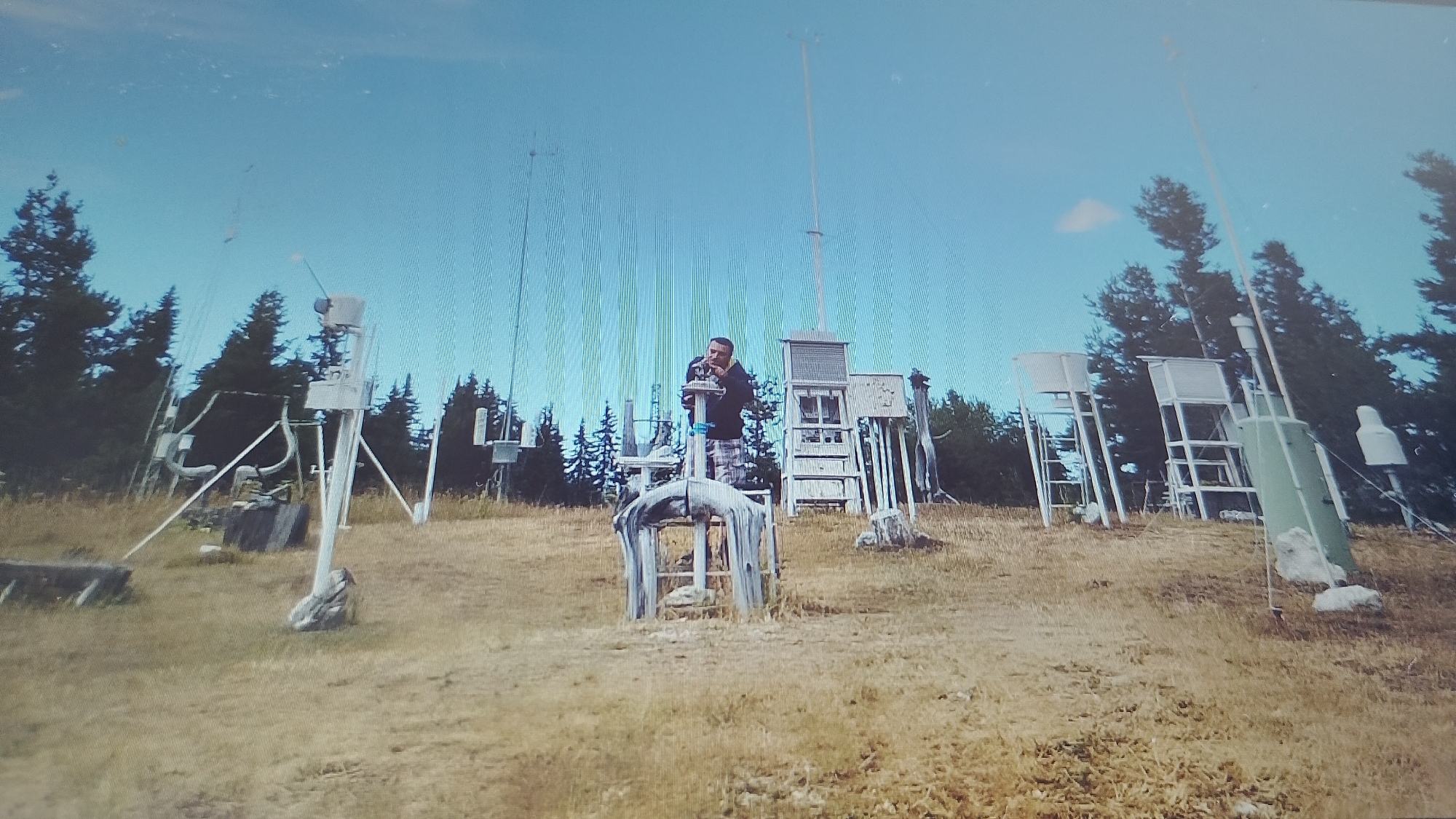 The image depicts a weather station situated in a grassy, hilly landscape with scattered trees in the background. A man, wearing a dark hooded sweatshirt and plaid pants, is standing on a platform, interacting with some of the weather monitoring equipment. The station consists of various structures and devices, including what appear to be anemometers, rain gauges, thermometers, and meteorological instruments housed in protective enclosures, mounted on poles and stands. The sky is clear and blue. This scene is consistent with a research or monitoring site, possibly located in the vicinity of Smolyan, Bulgaria, an area known for its mountainous terrain and climate research facilities.