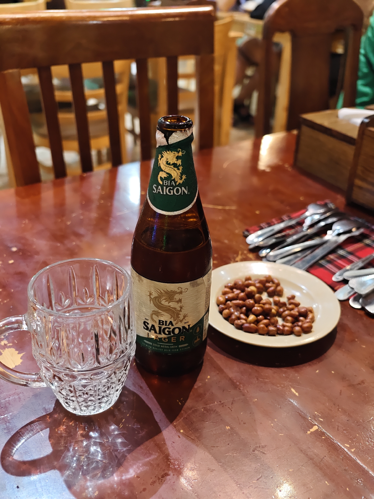 The image shows a close-up of a table at what appears to be an outdoor cafe or restaurant in Nha Trang Ward, Vietnam. The focus is on a bottle of Bia Saigon Lager, an empty glass mug with a patterned design, and a small plate of roasted peanuts. Spoons are neatly arranged on a red and black plaid napkin in the background, suggesting a meal or drinks are being served. Wooden chairs and parts of a wooden table are visible, along with hints of other patrons or staff in the blurred background, suggesting a lively atmosphere. The lighting suggests it could be daytime, possibly late afternoon or early evening. The overall mood seems relaxed and casual. The text "BIA SAIGON" is prominently displayed on the beer bottle, along with a dragon emblem and other smaller text indicating "LAGER" and "GOLD MEDAL BREW".