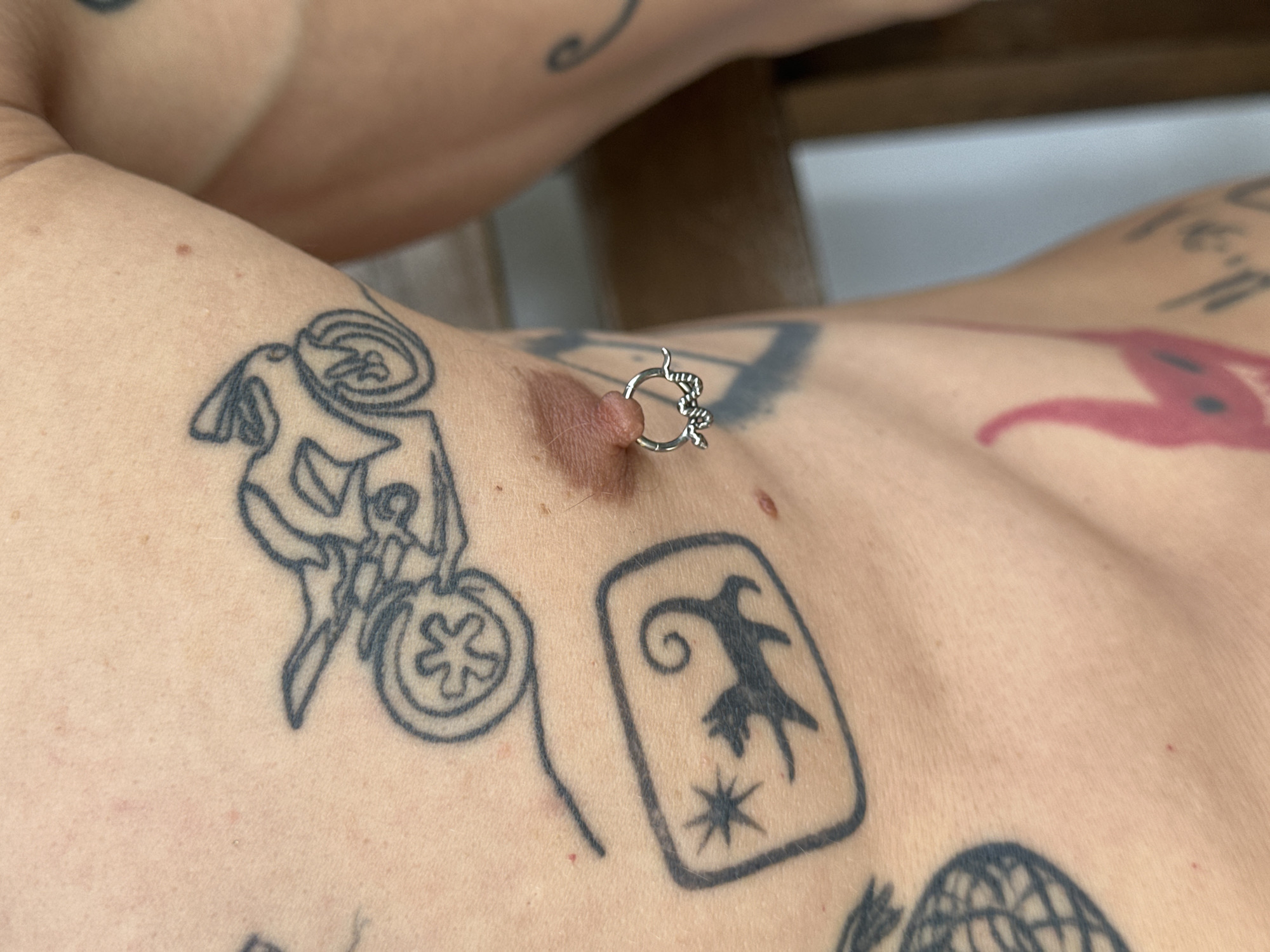 A close-up, eye-level shot focuses on the chest of a person with several tattoos and a nipple piercing. The person's skin is fair and appears to have some minor blemishes. Prominently featured is a tattoo of a motorcycle, drawn in a minimalist, outline style. To the left of the motorcycle tattoo is a rectangular design containing a stylized star and a silhouetted animal resembling a cat or fox with a curled tail. Below this, a portion of a pineapple tattoo is visible. The nipple piercing is a silver hoop with a snake design. The background is blurred, suggesting an indoor setting with wooden furniture. The image location is not discernible from the provided visual information.