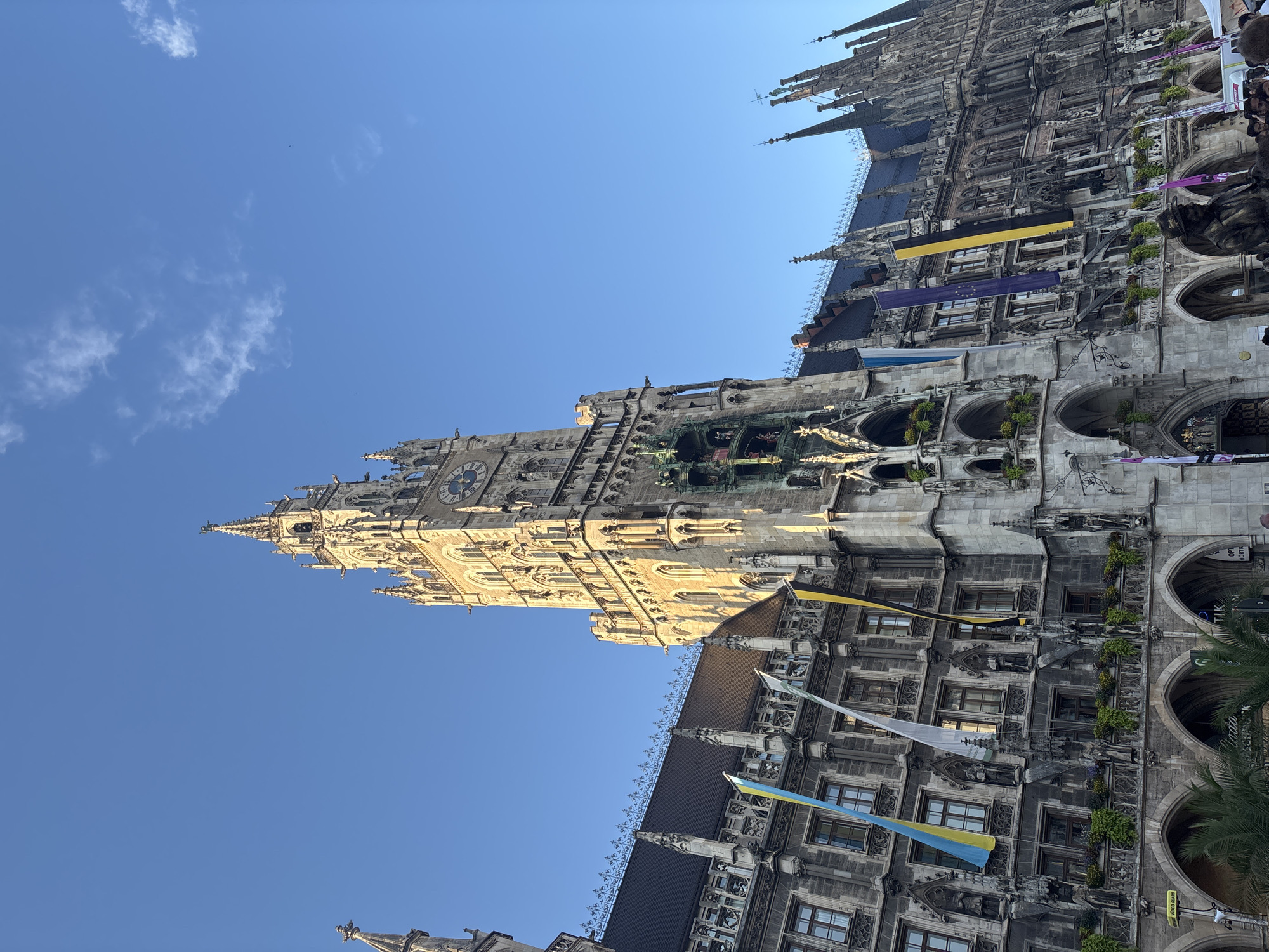 The image shows a detailed view of the New Town Hall (Neues Rathaus) located in Marienplatz, Munich, Germany. The grand Gothic Revival building features an ornate facade with numerous windows, decorative carvings, and arched walkways. A prominent clock tower rises majestically on the right side of the building. Several flags, including what appear to be Ukrainian and European Union flags, are hung vertically from the building's exterior. The sky above is a clear, bright blue with a few wispy clouds. Sunlight casts a warm glow on the stone structure, highlighting its architectural details. In the foreground, arches of the building are visible, and hints of people and stalls suggest a public square environment.