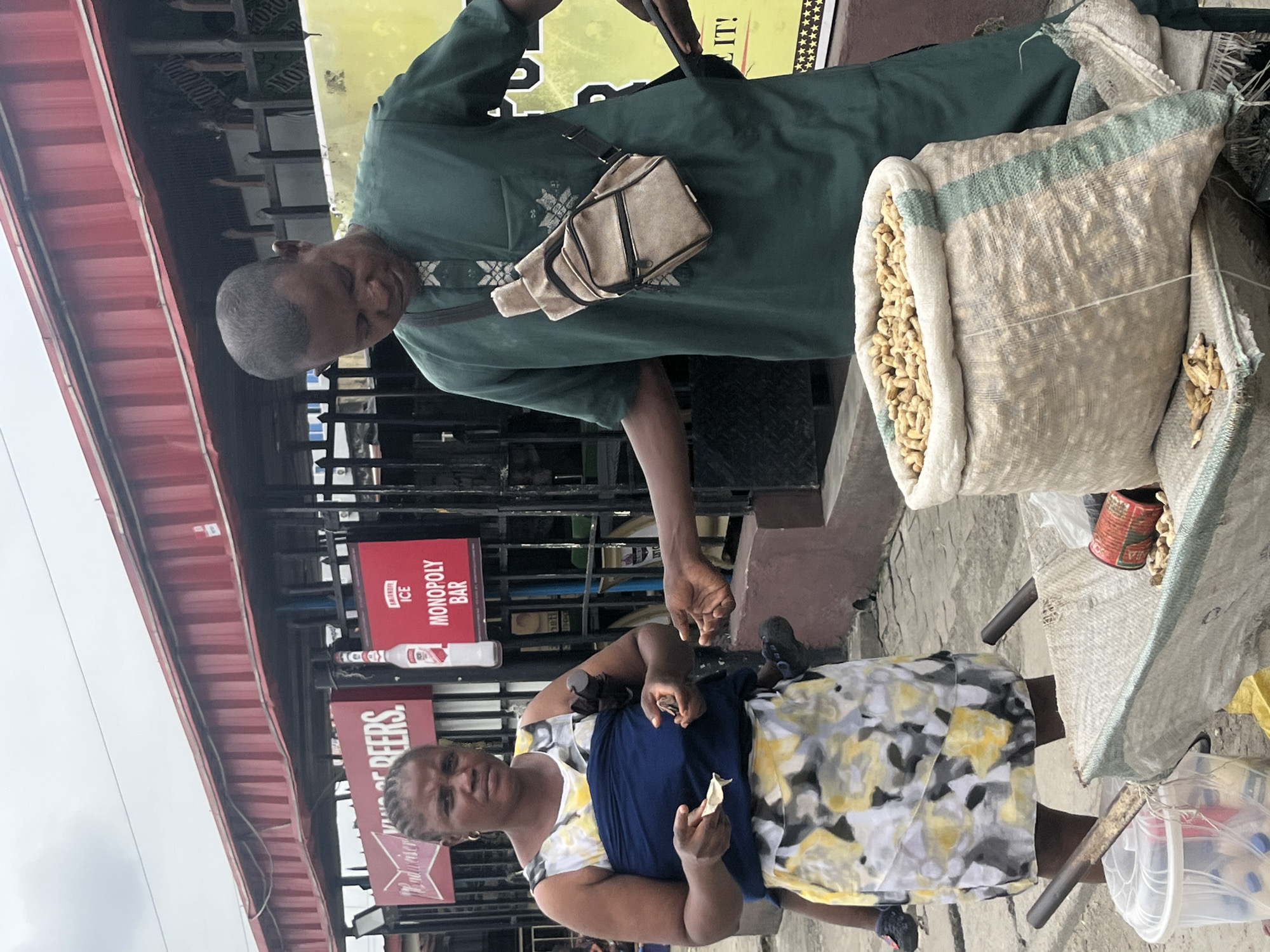 In Ado, Nigeria, a street vendor, a man in a green traditional outfit, stands by a large sack of peanuts. He is looking down at his hand, which is extended towards another person's hand. A woman, wearing a yellow and white floral dress over a blue baby carrier, faces the man and holds a small amount of money in her hand. A cart with a clear plastic container of bottled drinks and a red can is positioned in front of them. Behind the individuals, a red awning stretches across the scene, with signs for "Budweiser Beers" and "Monopoly Bar" visible. The ground is paved with concrete, and a dark metal fence runs along the background.