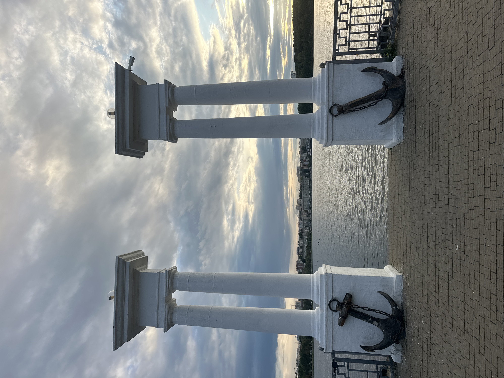 The image shows a waterfront promenade with two classical white columns framing a view of a lake or river. Anchors are affixed to the base of each column. The sky is cloudy with hints of sunlight breaking through, suggesting either sunrise or sunset. In the distance, a city skyline is visible along the opposite shore, characterized by buildings and trees. A metal railing runs along the edge of the promenade. The ground is paved with brickwork. There are no people visible in the image. This scene is located at the Ternopil Lake embankment in Ternopil, Ukraine.