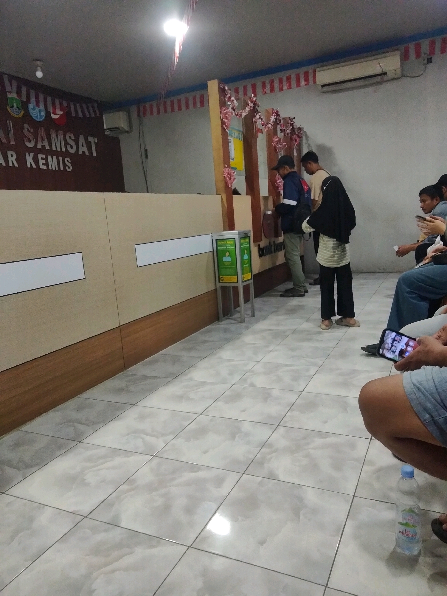 The image depicts the interior of a government office, likely a Samsat office (vehicle registration and identification office) in Sukaasih, Indonesia. The scene is brightly lit, with fluorescent lights on the ceiling. The walls are painted a light gray, and the floor is tiled with large, light-colored tiles that have a marble-like pattern.
In the background, a counter with a beige and wood-grain finish spans across the room. Above the counter, a dark brown sign reads "AI SAMSAT" and "AR KEMIS". Red and white striped decorations and small pink flowers are visible along the top of the wall and around a wooden pillar. An air conditioning unit is mounted on the wall.
Several individuals are present in the office. In the middle ground, two men stand near the counter. One man, wearing a blue jacket and a cap, appears to be interacting with a machine or a service window. The other man, in a light-colored shirt and dark pants, is standing next to him. Further to the right, a person wearing a black hijab and a striped shirt stands, facing away from the camera.
In the foreground, several people are seated and waiting. One individual, partially visible on the right, is wearing blue shorts and is holding a smartphone. Another person, seated on the far right, is wearing a light blue shirt and is looking at their phone. A bottle of "Le Minerale" water is placed on the tiled floor in the foreground. A dispenser with green signage is positioned next to the counter. The overall atmosphere suggests a typical public service waiting area.