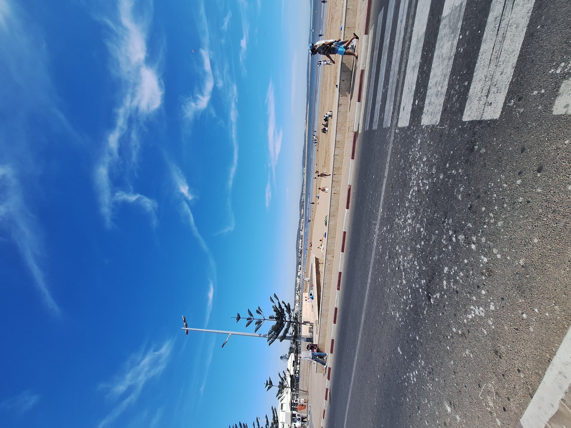 The image depicts a view of the Essaouira beachfront and promenade under a bright blue sky with wispy clouds. In the foreground, a paved road with a pedestrian crossing is visible, showing some splatter marks. Along the edge of the road, a concrete curb separates it from a paved walkway. Beyond the walkway, a sandy beach stretches out, populated by several individuals and small groups. Some people are sitting or lying on the sand, while others appear to be walking or standing. The ocean is visible in the background, with a distant coastline and buildings. A lamppost with multiple lights stands prominently on the promenade, and several trees with distinctive shapes are planted along the walkway. The scene suggests a leisurely day at the beach, with people enjoying the coastal environment.