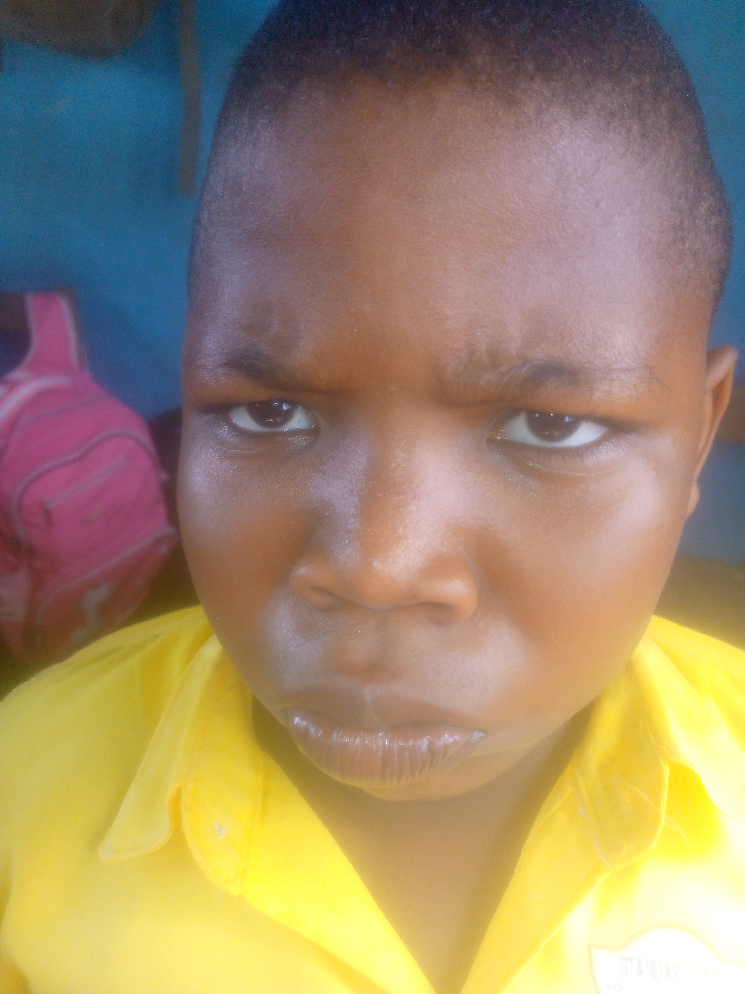 A close-up photograph shows a young, dark-skinned person with a closely shaved haircut. The person is wearing a yellow shirt with a buttoned collar; a pink backpack is partially visible in the background. The person has a furrowed brow and appears to be frowning, suggesting a mood of seriousness or displeasure. The photograph's origin is suggested to be Ikorodu, Nigeria, but the image provides no verification.